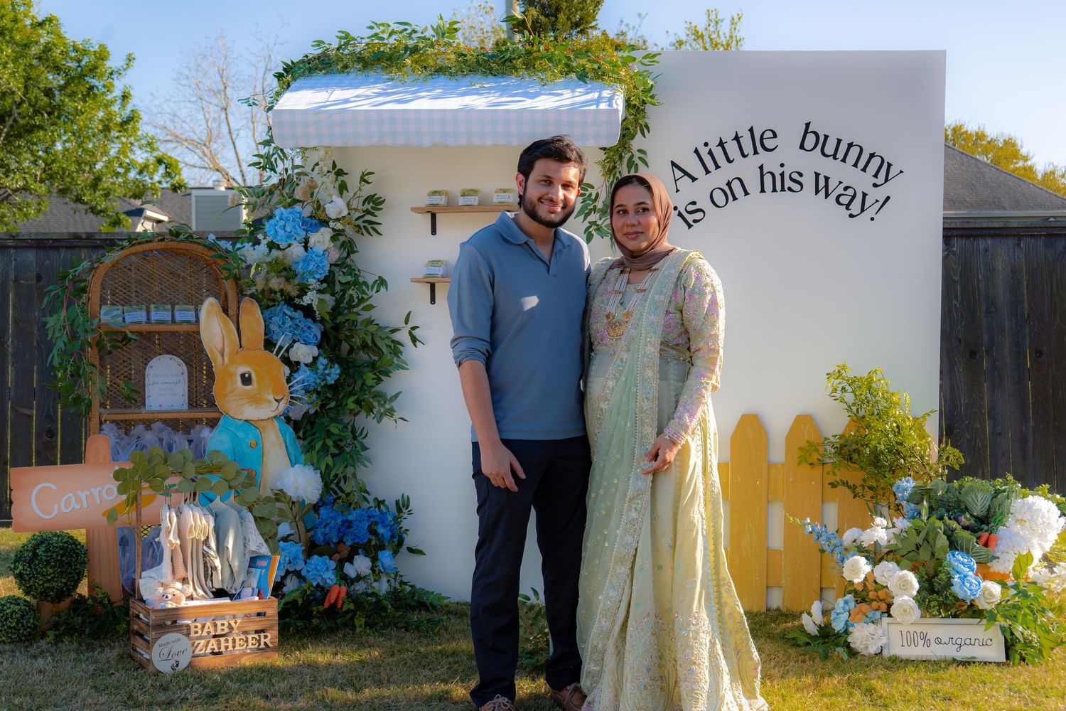 Expectant couple smiling at Peter Rabbit baby shower with blue and green garden party decorations.