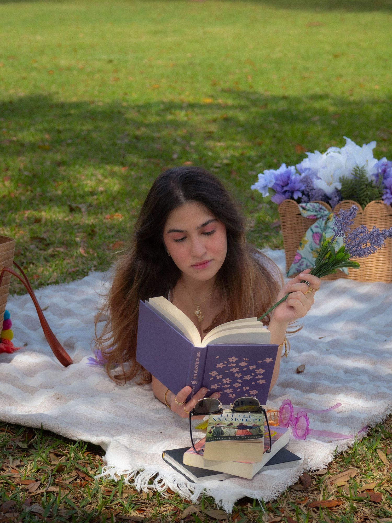 Young woman lying on picnic blanket in park reading a purple book while holding blue wildflowers.