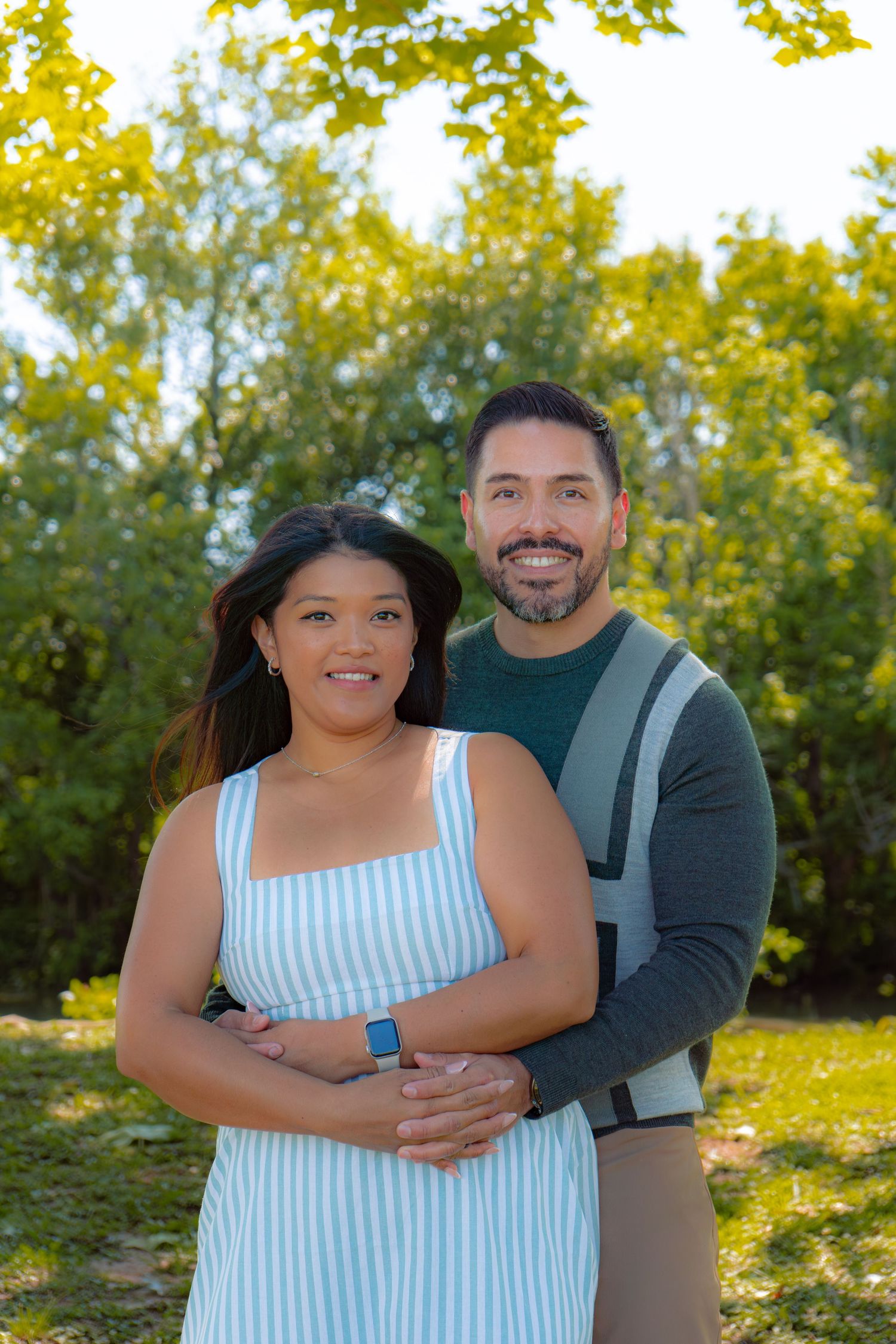 A smiling couple poses outdoors in a lush green park, woman in striped dress, man in green sweater embracing her.
