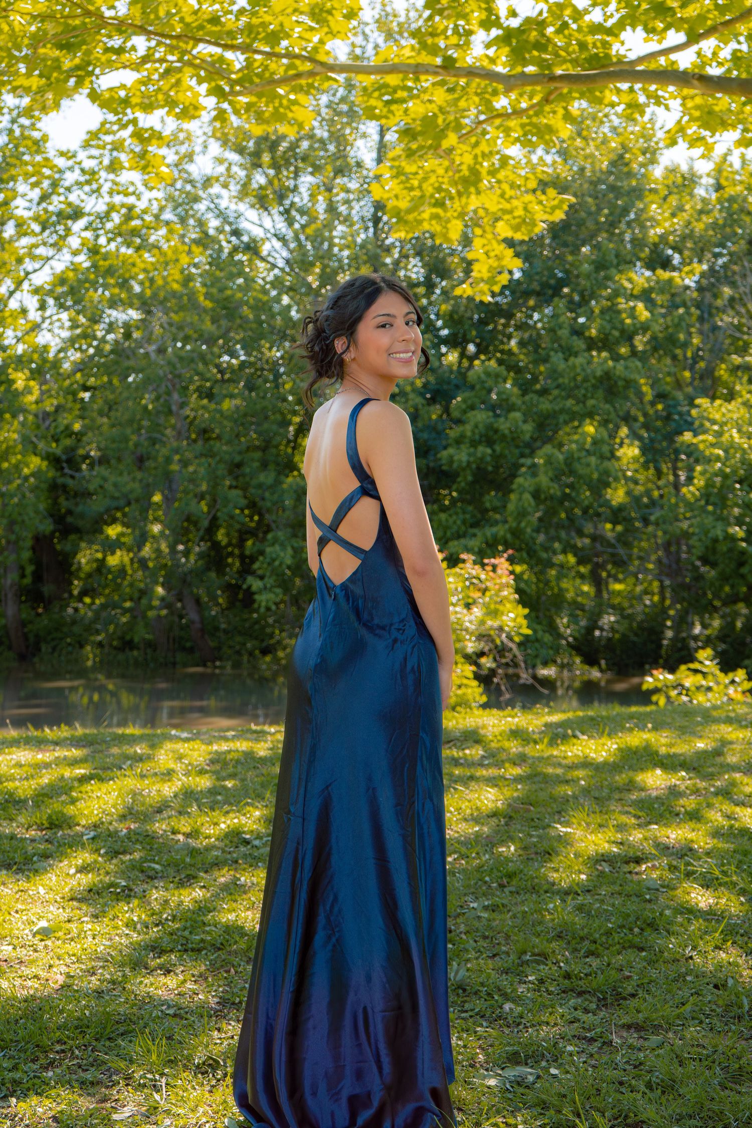 Young woman in elegant navy blue backless satin gown smiling outdoors among lush green trees.