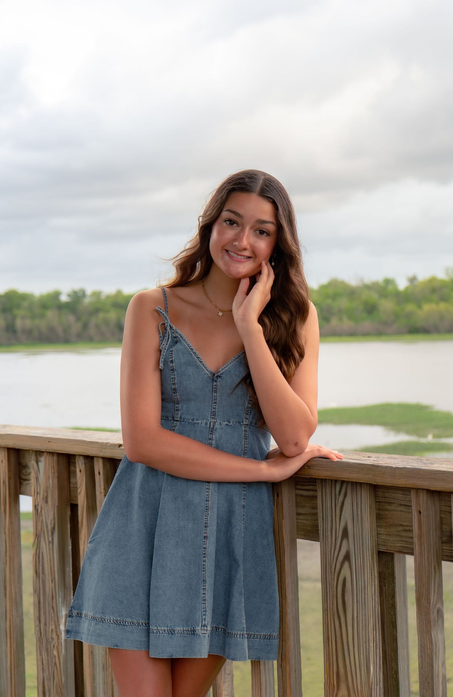 Young woman in denim mini dress posing on wooden deck overlooking scenic waterway.