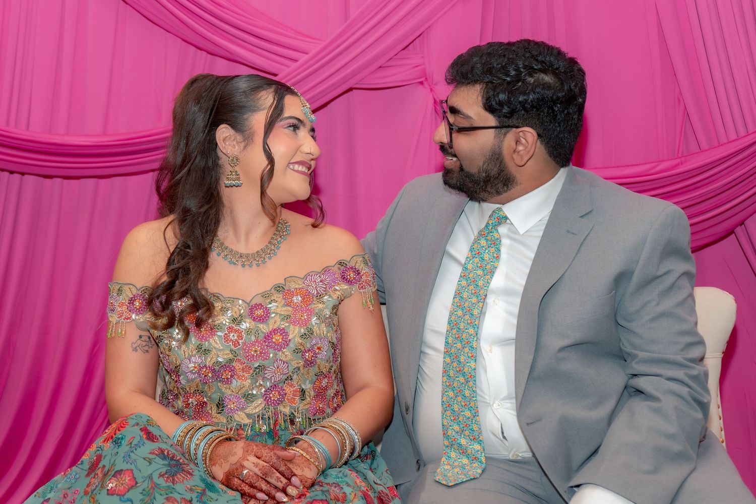Couple in traditional Indian attire smiling at each other at a festive celebration with pink backdrop.