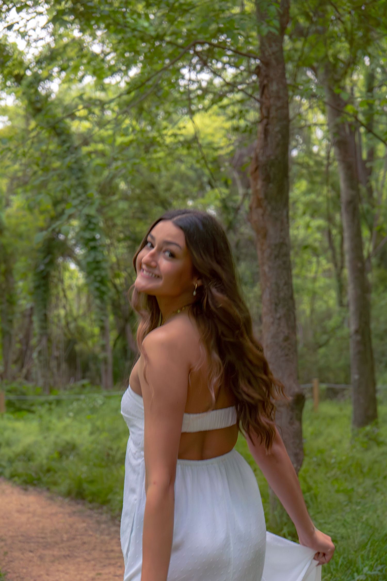 Smiling young woman in white dress posing on a forest trail surrounded by trees.