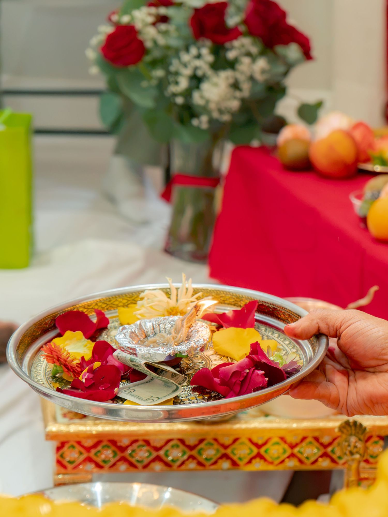 Hand holding a silver puja thali with rose petals, diya, and offerings at a Hindu ceremony.