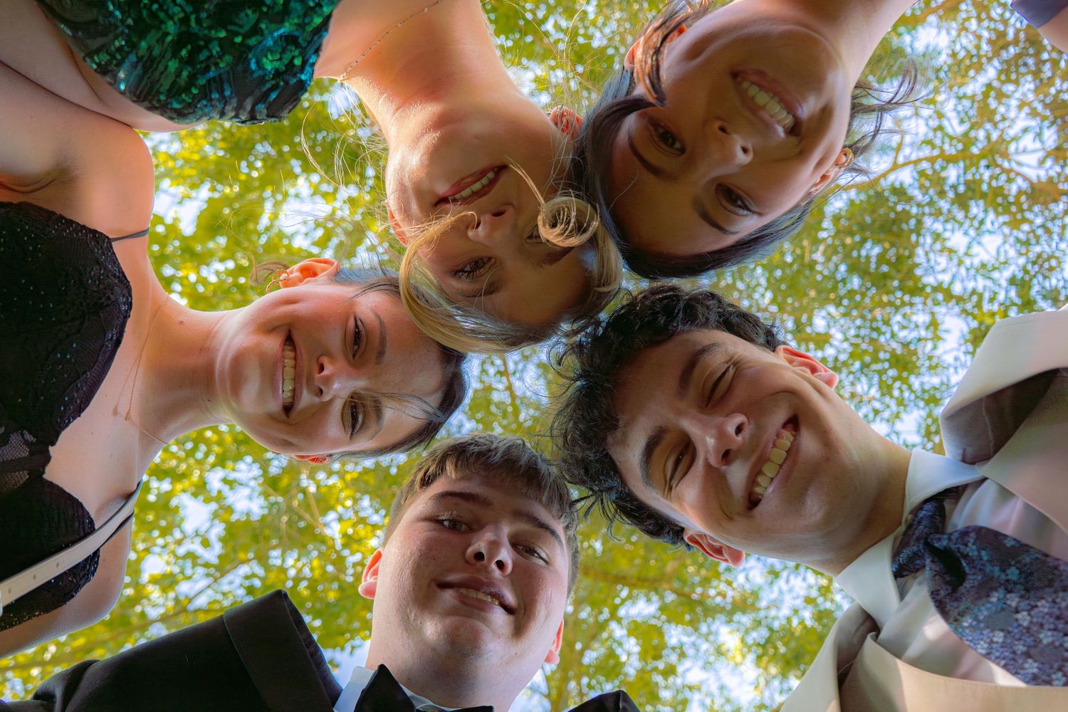 Five smiling teens huddle together in a circle looking down at camera with green trees overhead.
