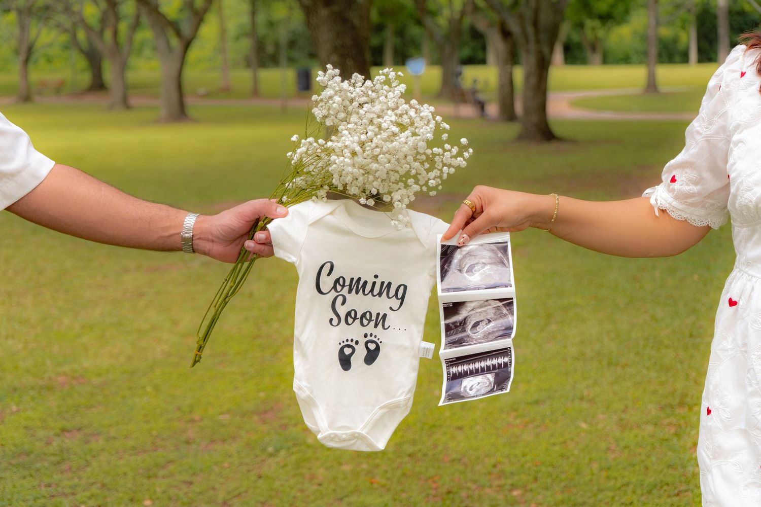 Couple holding a white Coming Soon baby onesie with tiny footprints and baby's breath flowers for pregnancy announcement.