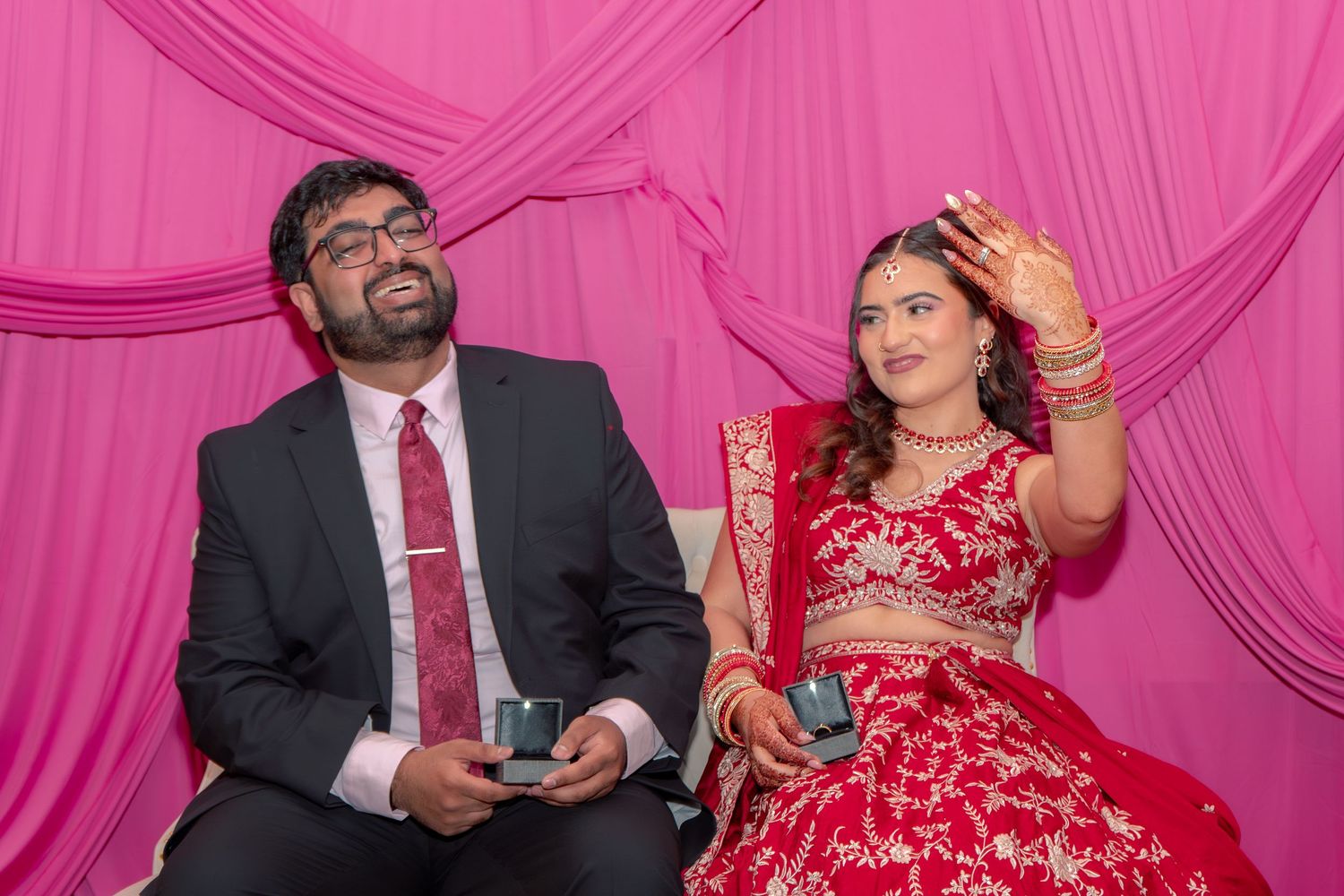 South Asian couple seated before pink drape, bride in red lehenga waving hand while groom in black suit looks up smiling.