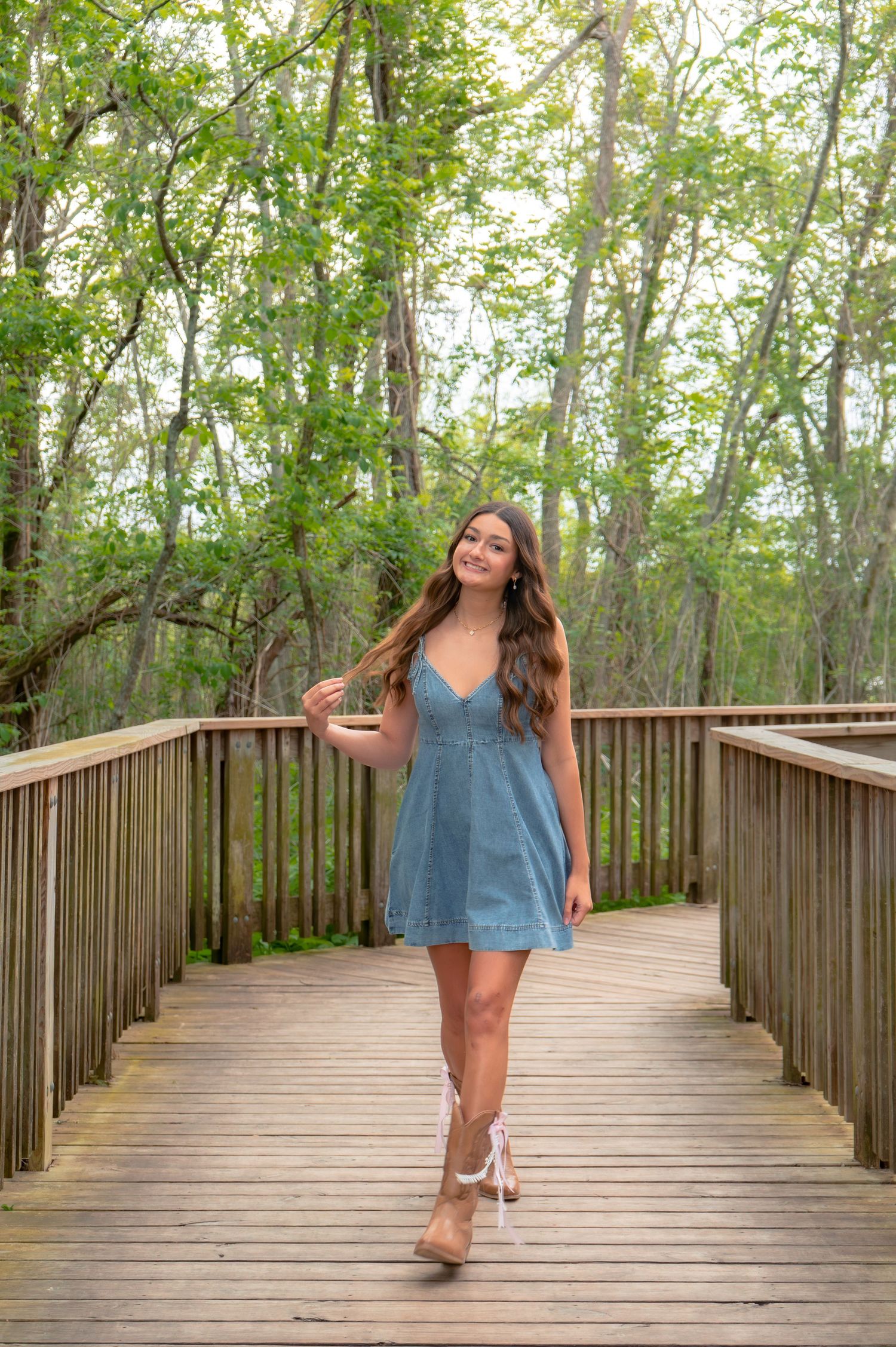 Young woman in denim dress and lace heels walking on wooden boardwalk surrounded by lush green trees.