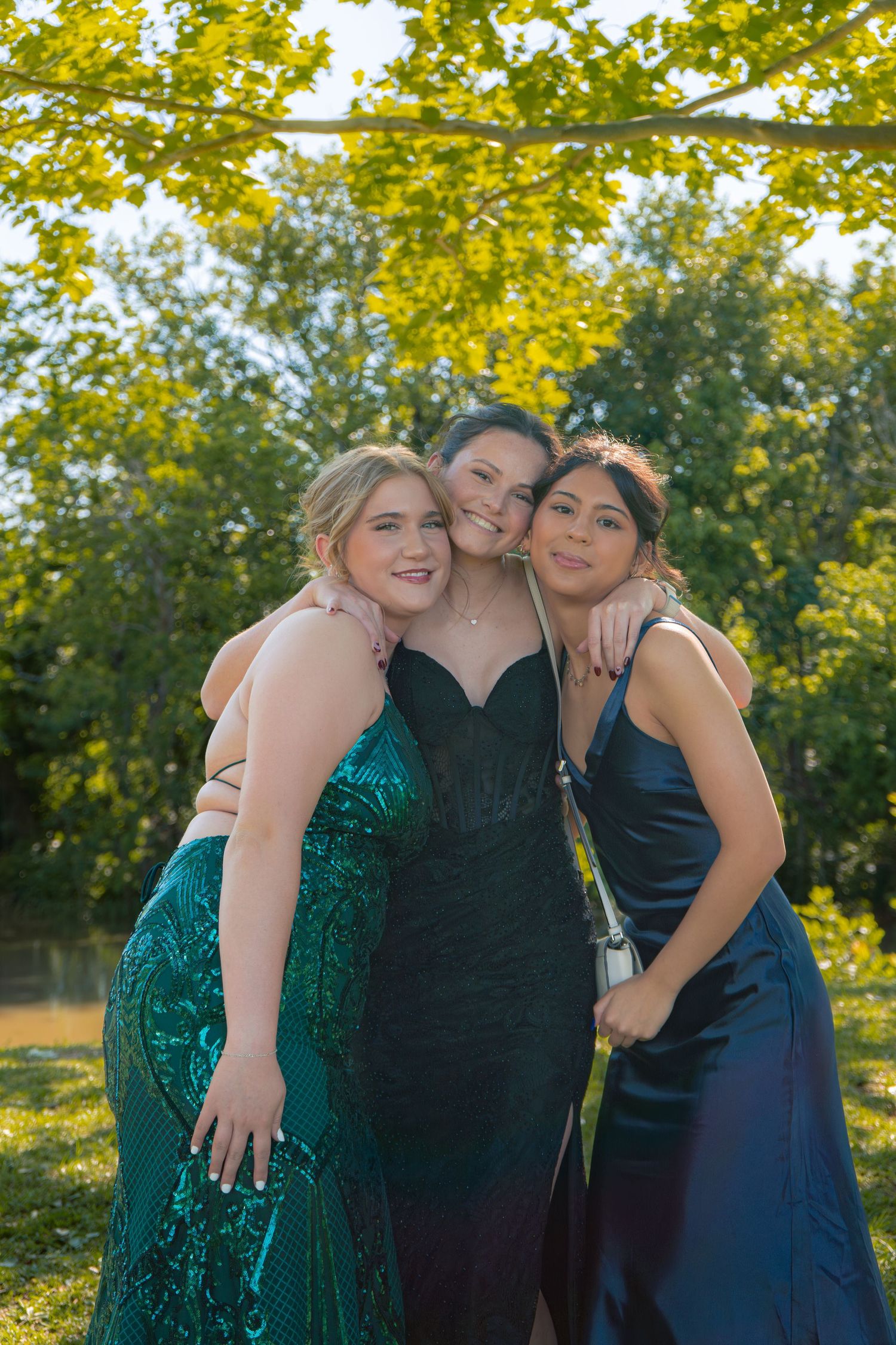 Three smiling teenage girls in formal prom dresses embrace outdoors under golden sunlit tree branches.