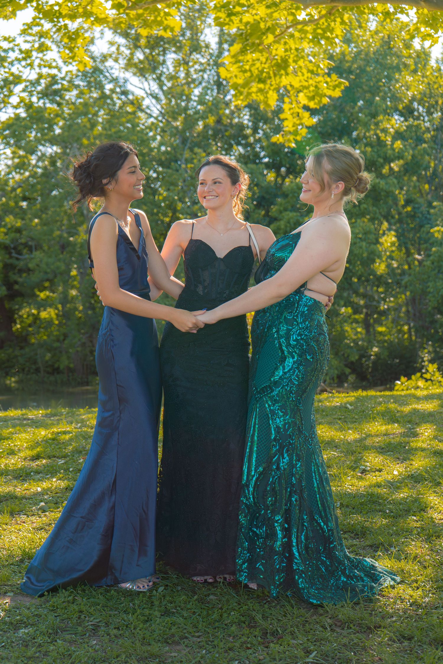 Three young women in elegant prom gowns hold hands and smile in a sunlit park.