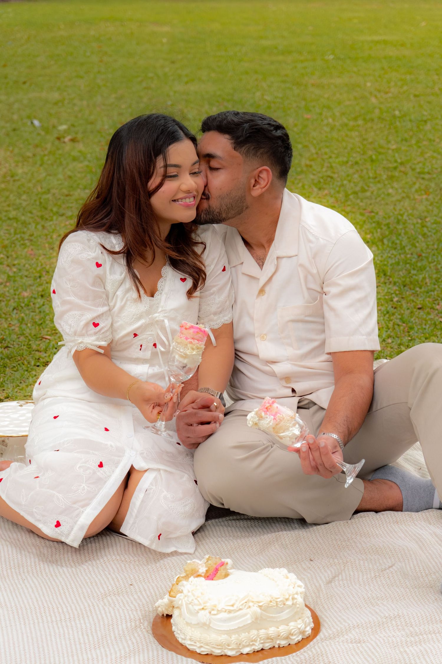 Happy couple sitting on grass sharing champagne glasses and kissing beside a white heart-shaped cake.