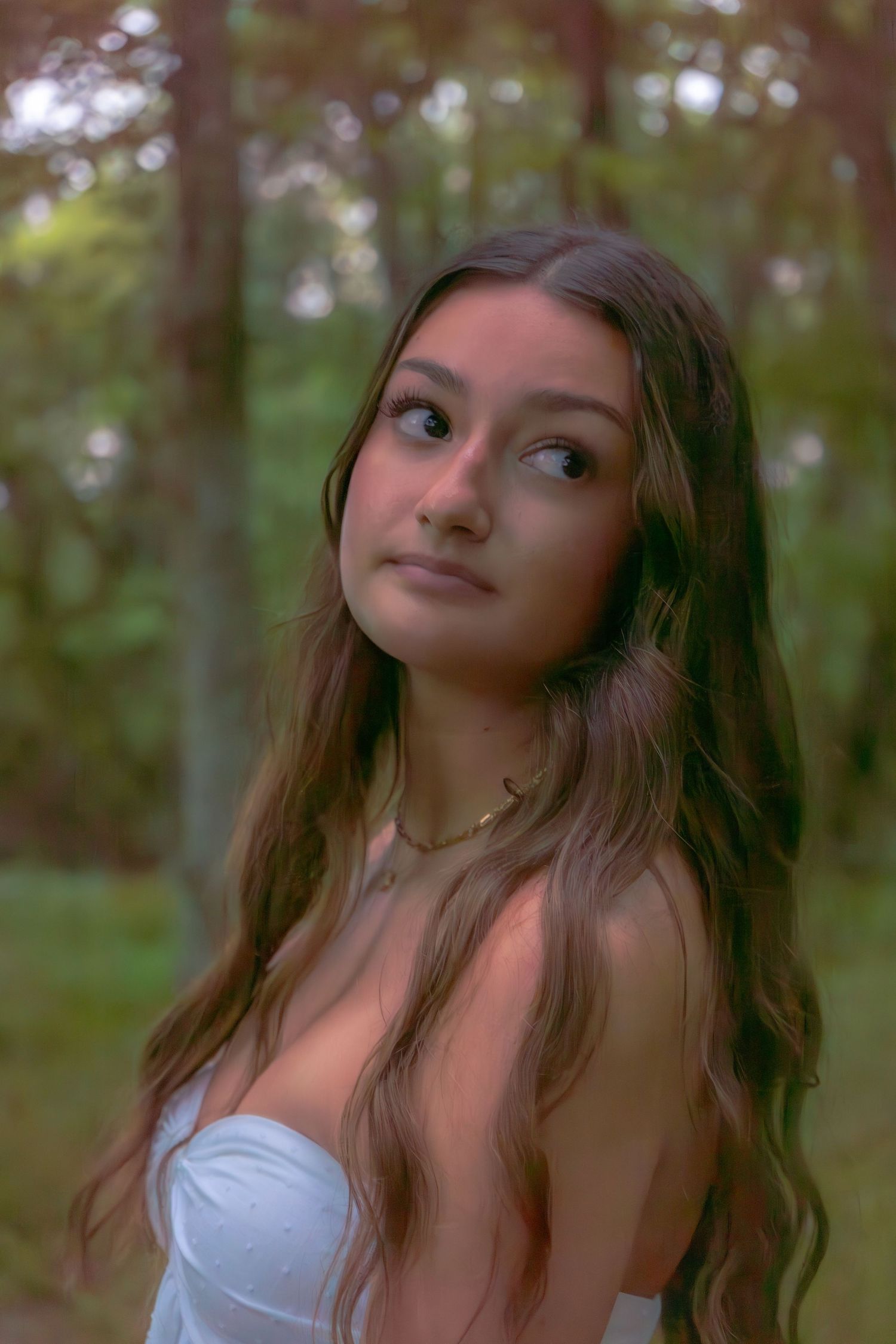 Young woman with long wavy hair wearing white strapless top and gold necklace in forest.