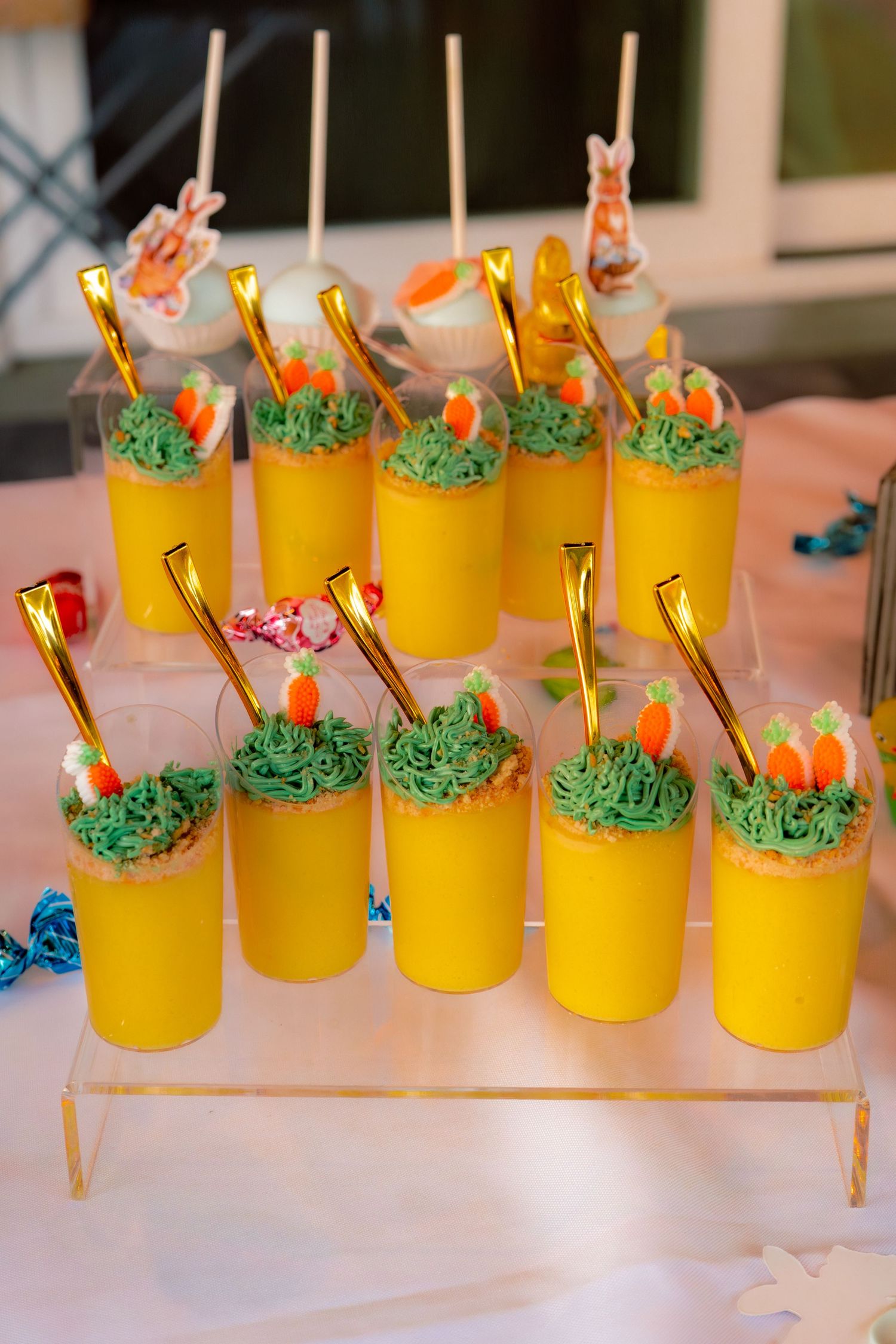 Yellow dessert cups with green frosting, carrot decorations, and gold spoons displayed on a clear acrylic stand.