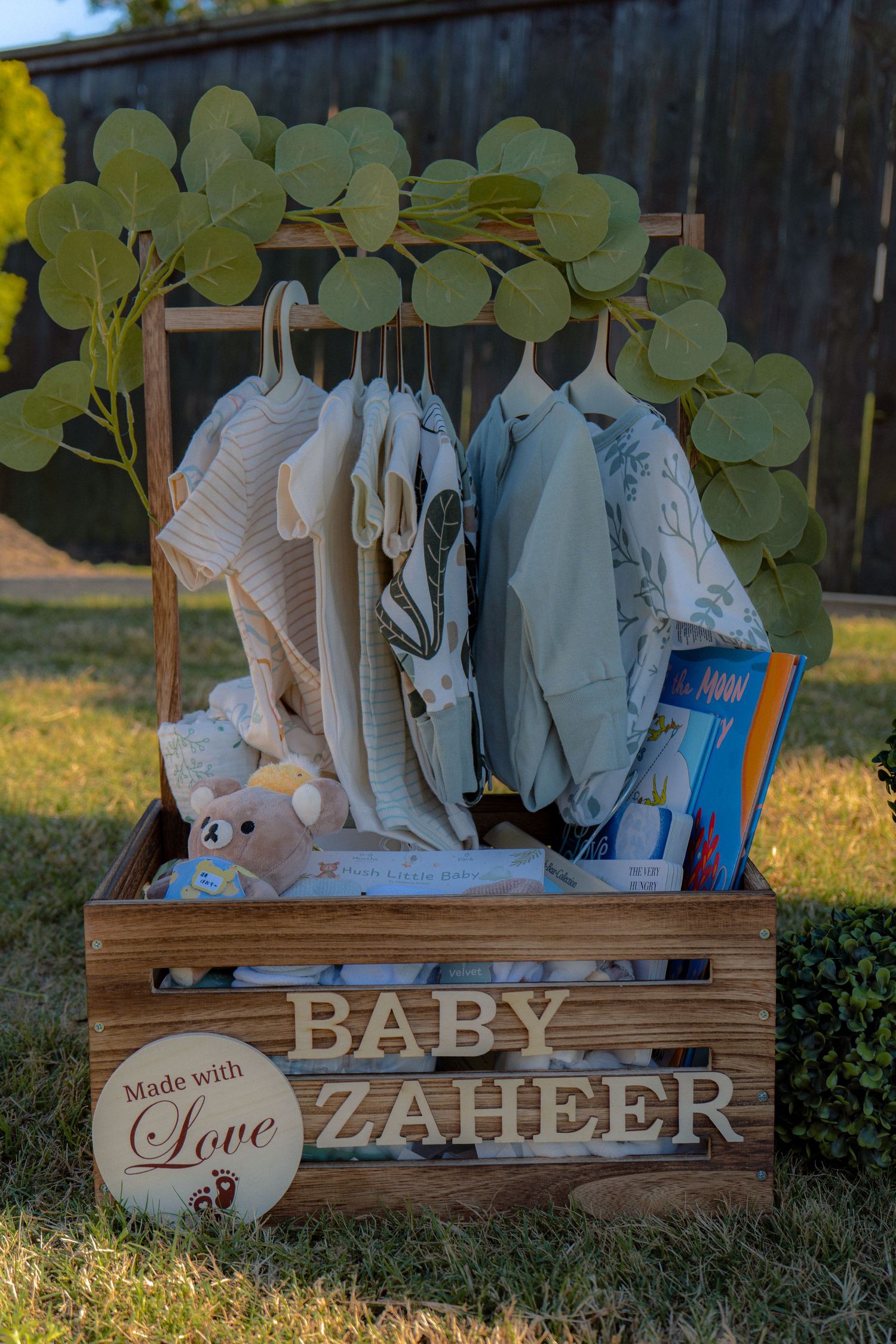 Wooden crate baby shower gift display named Baby Zaheer with hanging infant clothes and eucalyptus decor outdoors.