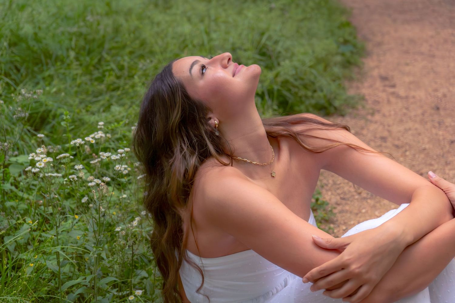Young woman in white dress sitting in lush green grass, looking up serenely with long brown hair flowing.