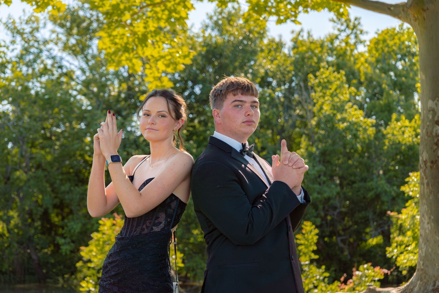 A teenage couple in formal prom attire pose back-to-back in a James Bond-style stance outdoors among green trees.