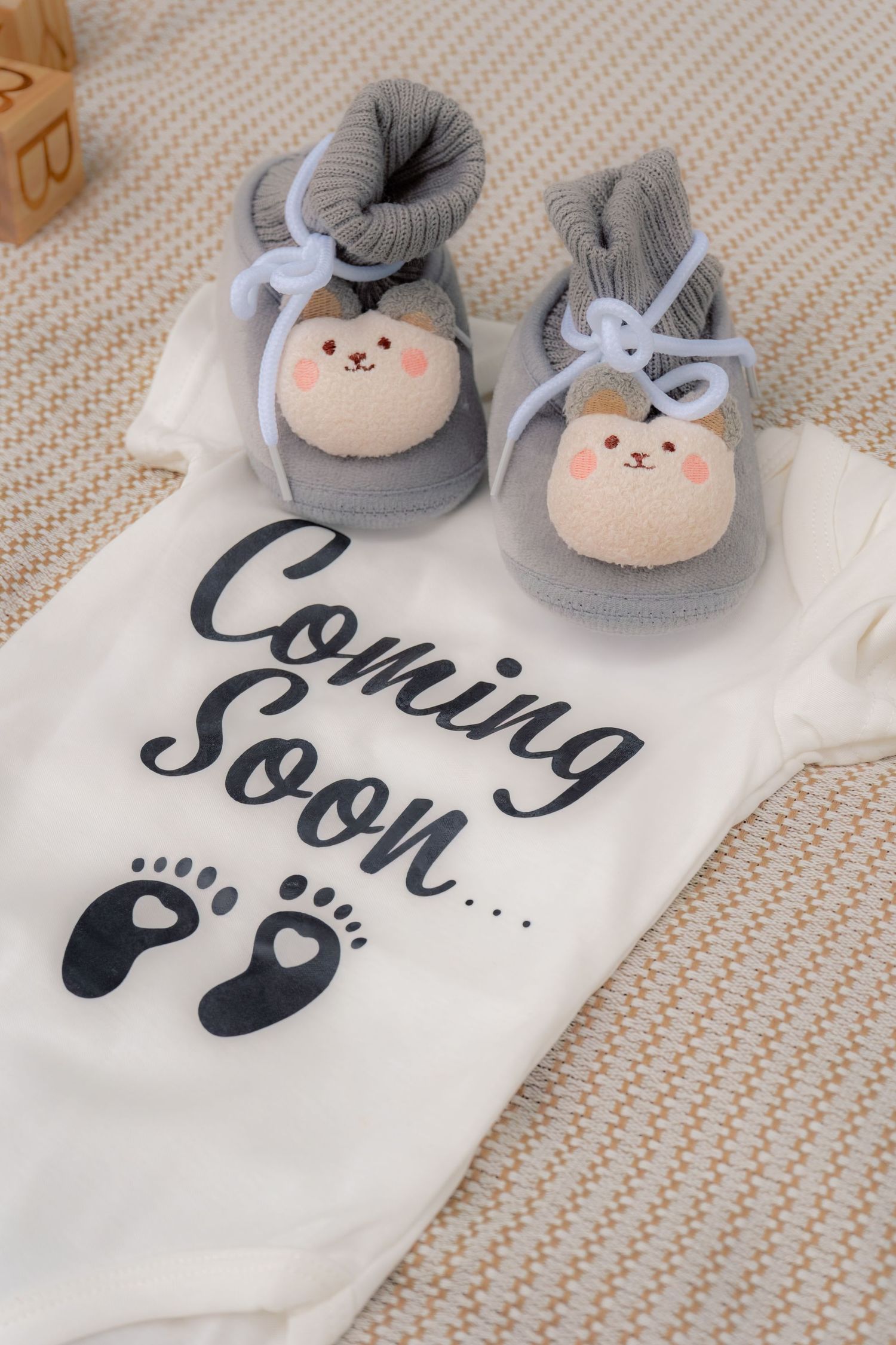 White baby onesie with Coming Soon text and tiny footprints beside cute gray bear booties on woven mat.