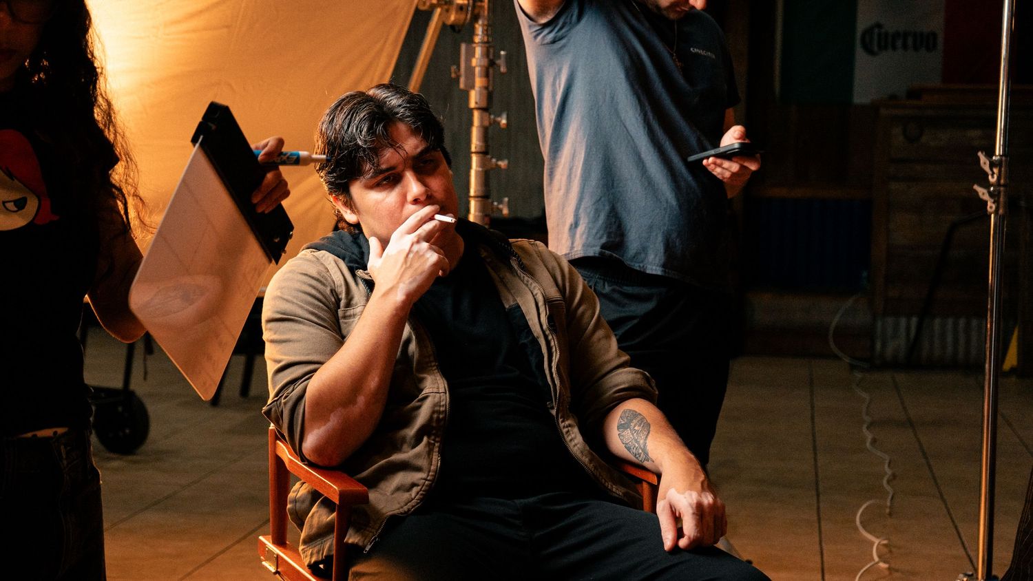 A tattooed man sits in a director's chair on a film set, thoughtfully touching his face.