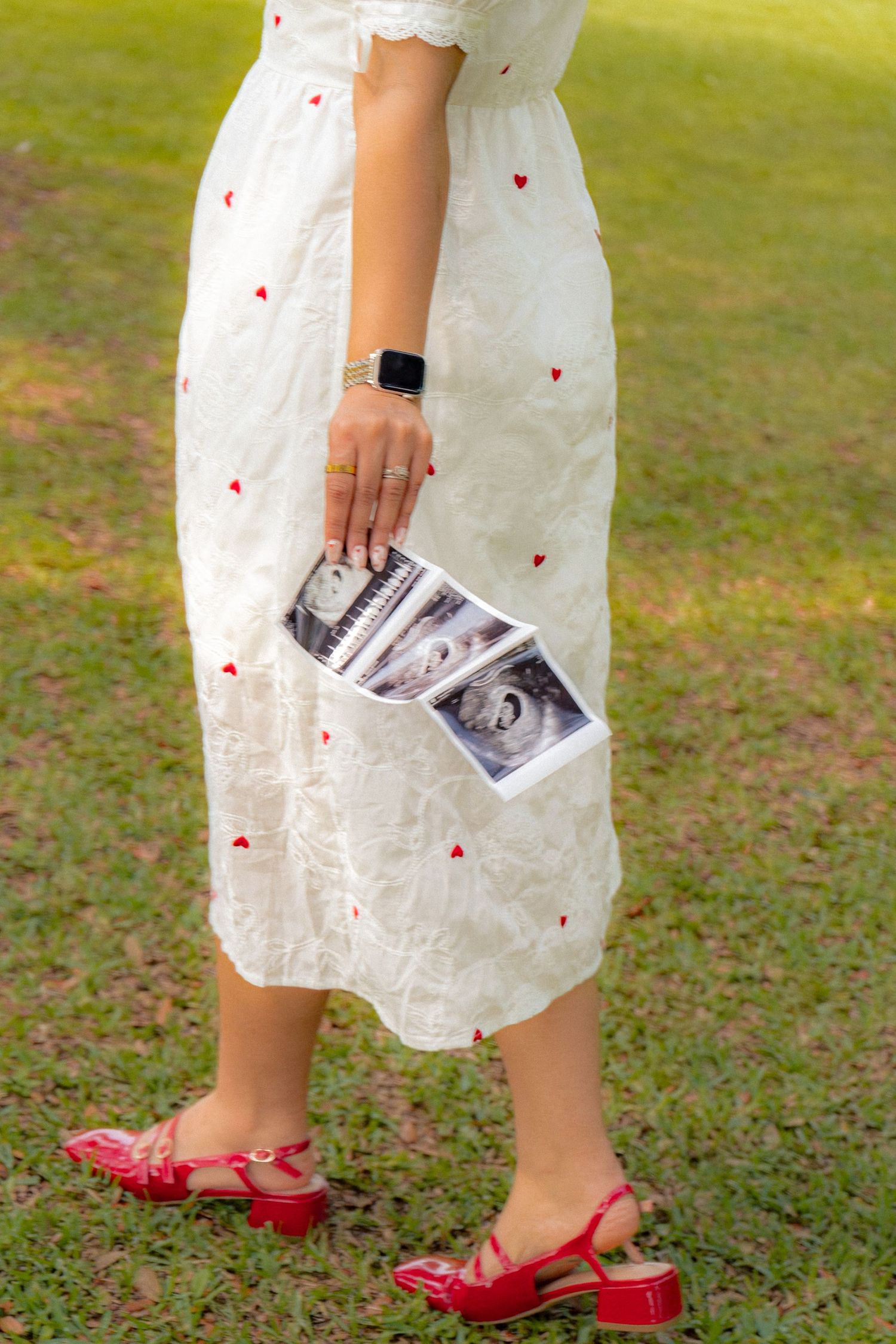 Pregnant woman in white heart-print dress and red heels holding ultrasound photos outdoors.