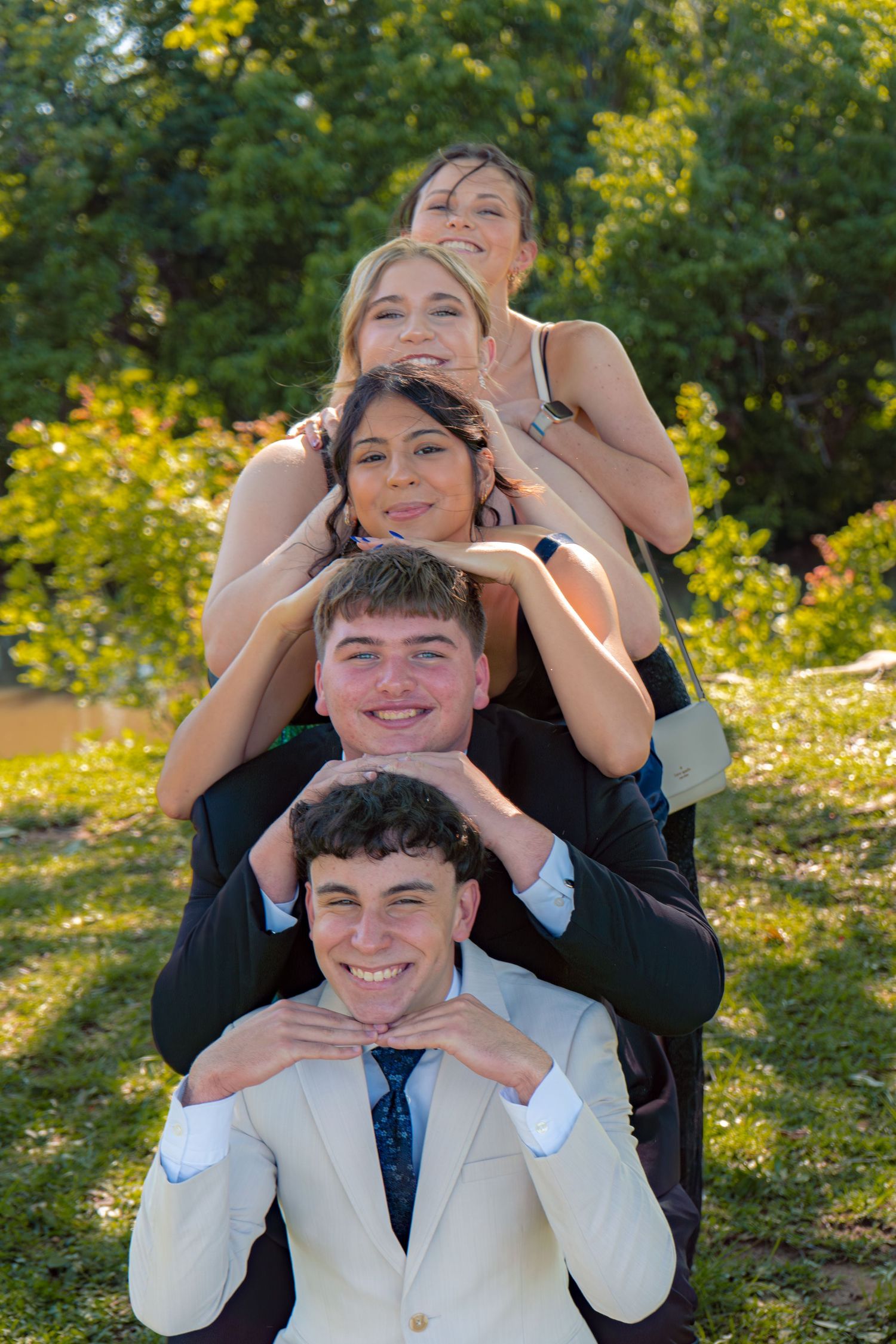 Five prom-dressed teens pose in a fun stacked formation outdoors in a sunny green park.