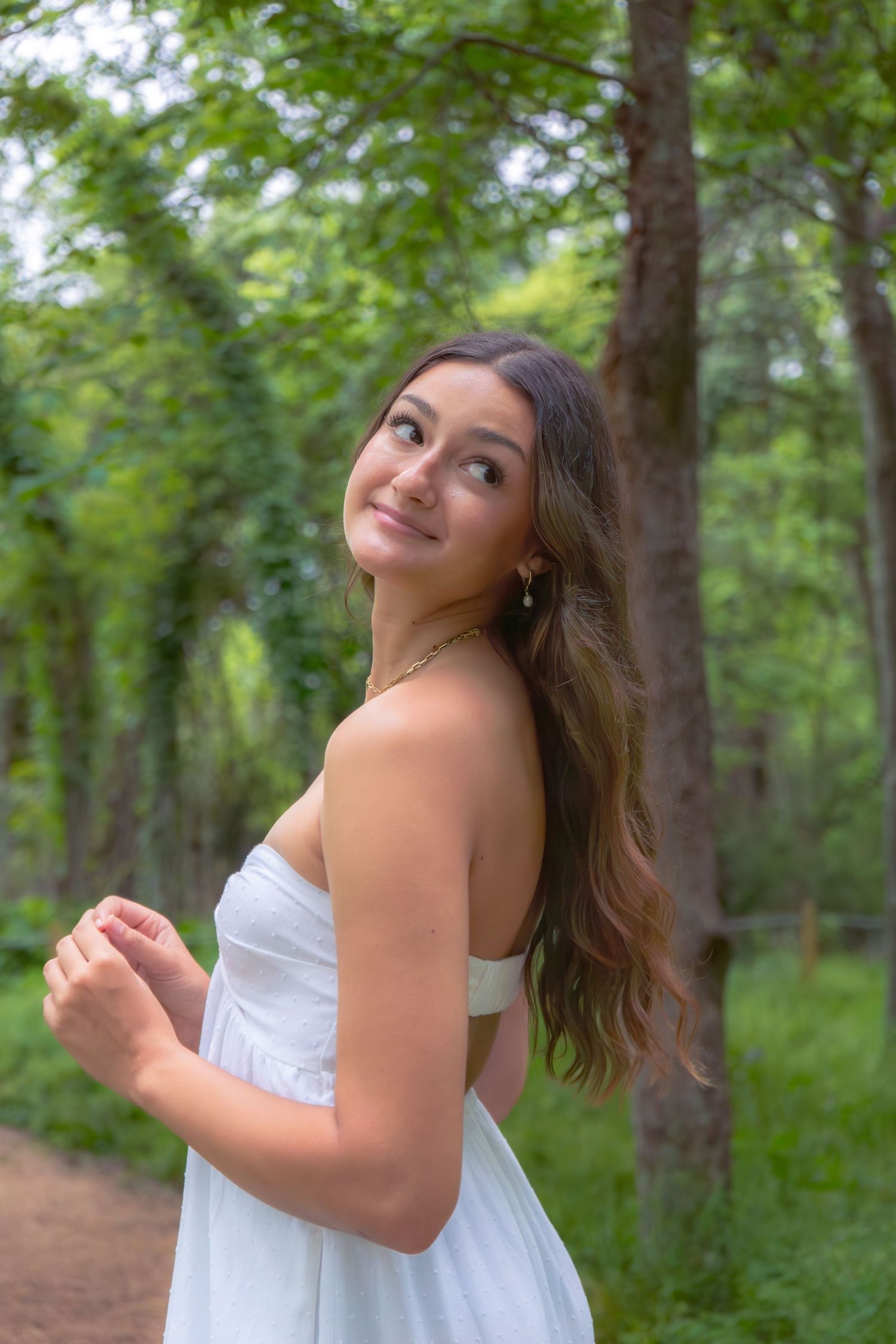 Young woman in white strapless dress smiling over shoulder on forest path surrounded by lush green trees.