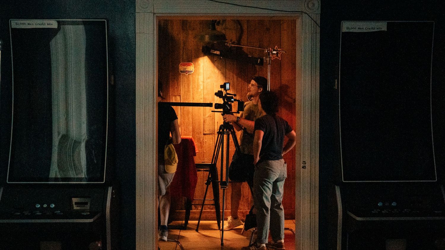 Film crew operating a camera on a tripod inside a dimly lit room during a production shoot.