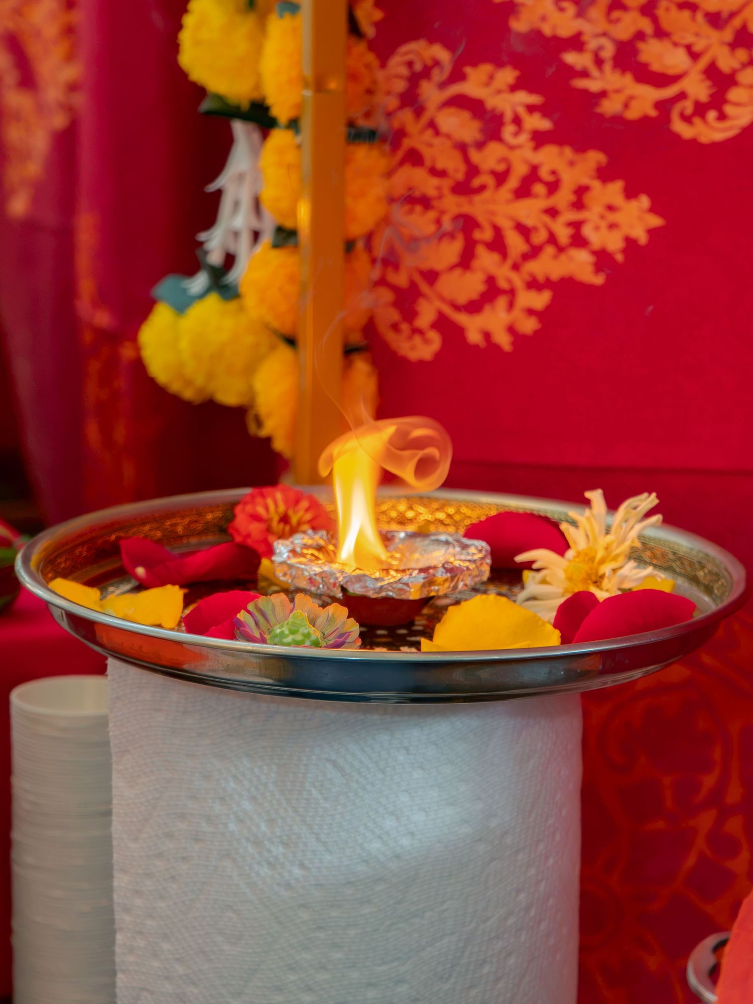 A lit diya on a silver puja thali with colorful flowers, set against a vibrant red backdrop with marigold garlands.