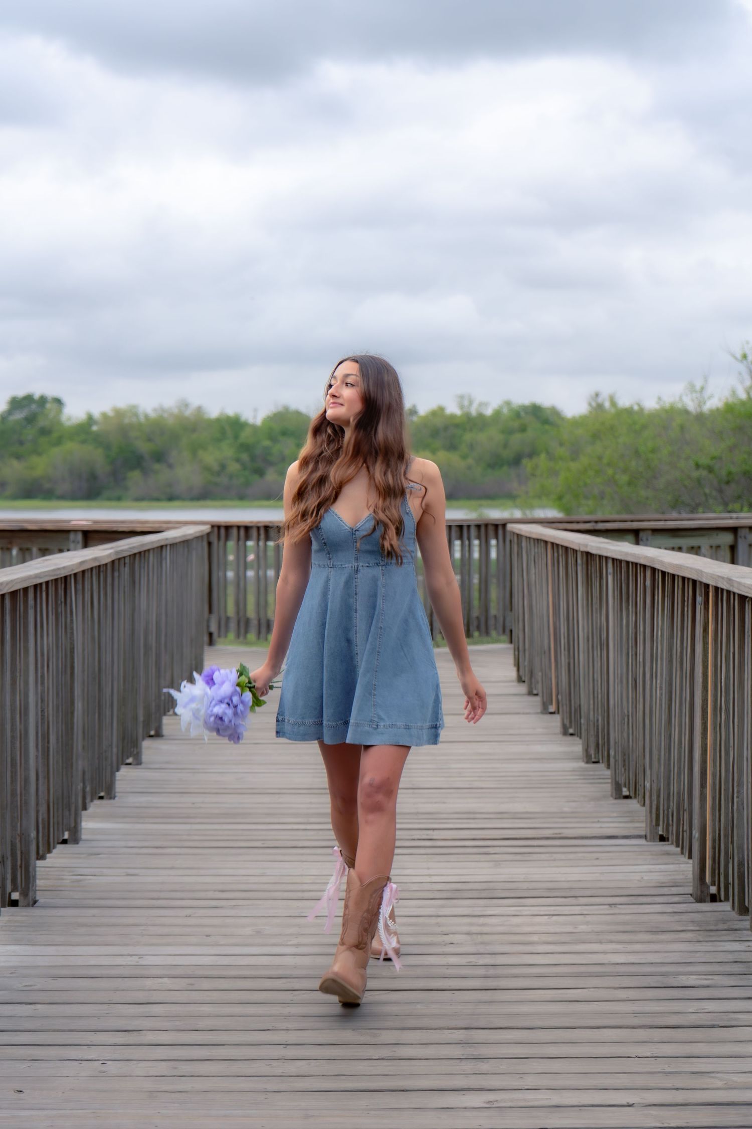 Young woman in denim dress holding purple flowers walks on wooden bridge boardwalk on cloudy day.