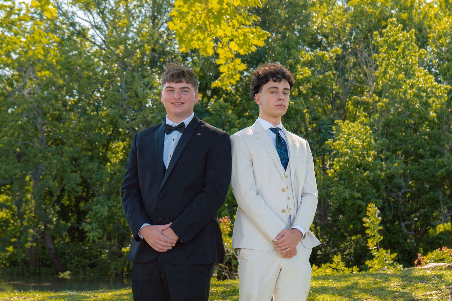 Two young men in formal suits posing outdoors at prom, one in black tuxedo and one in cream suit.