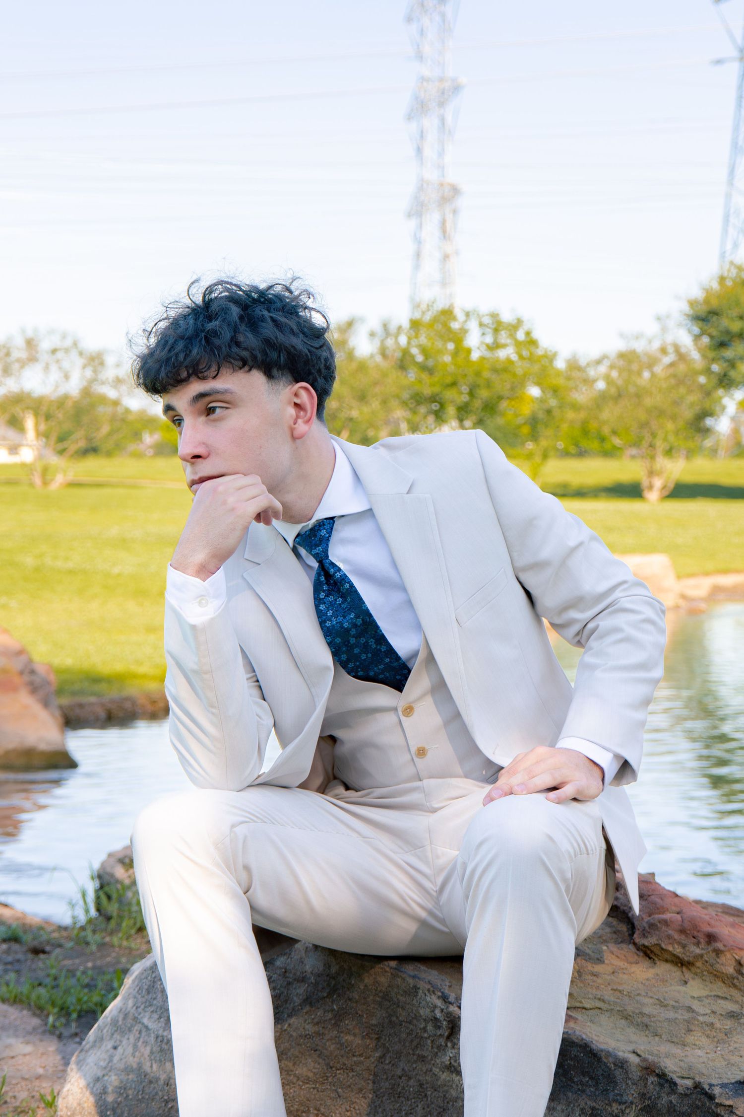 Young man in white suit and blue tie sits on rocks by a pond, gazing thoughtfully into the distance outdoors.