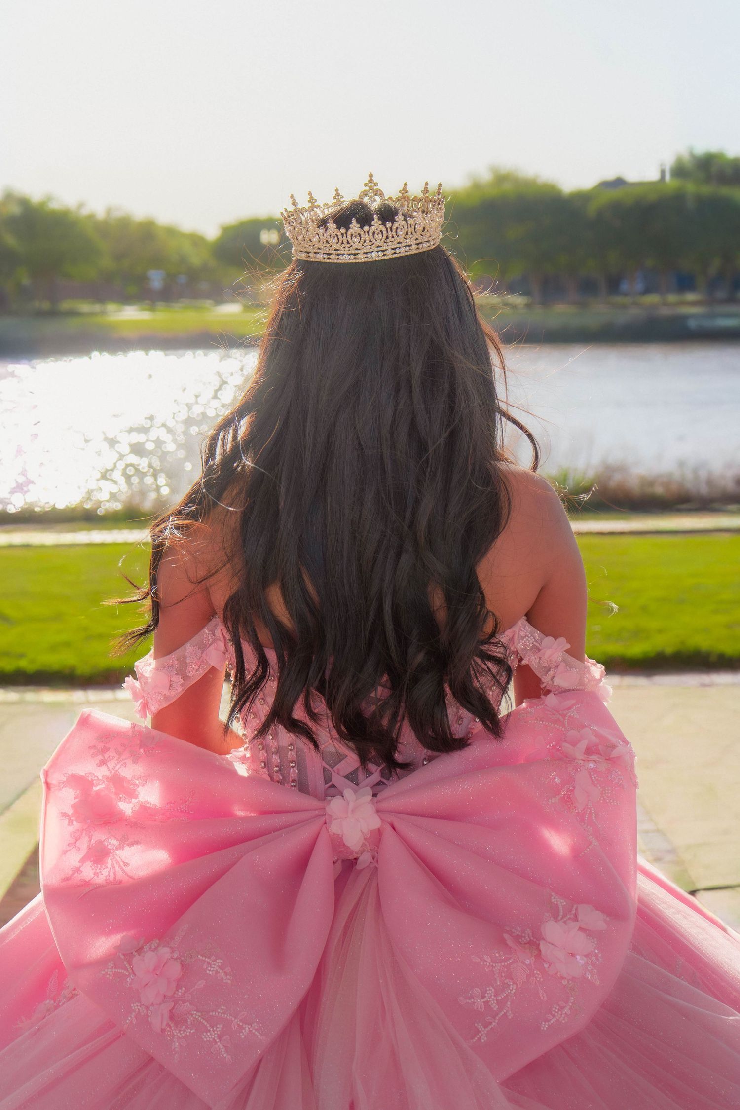 Young woman in pink ball gown with gold crown and large bow, viewed from behind near a serene lake.