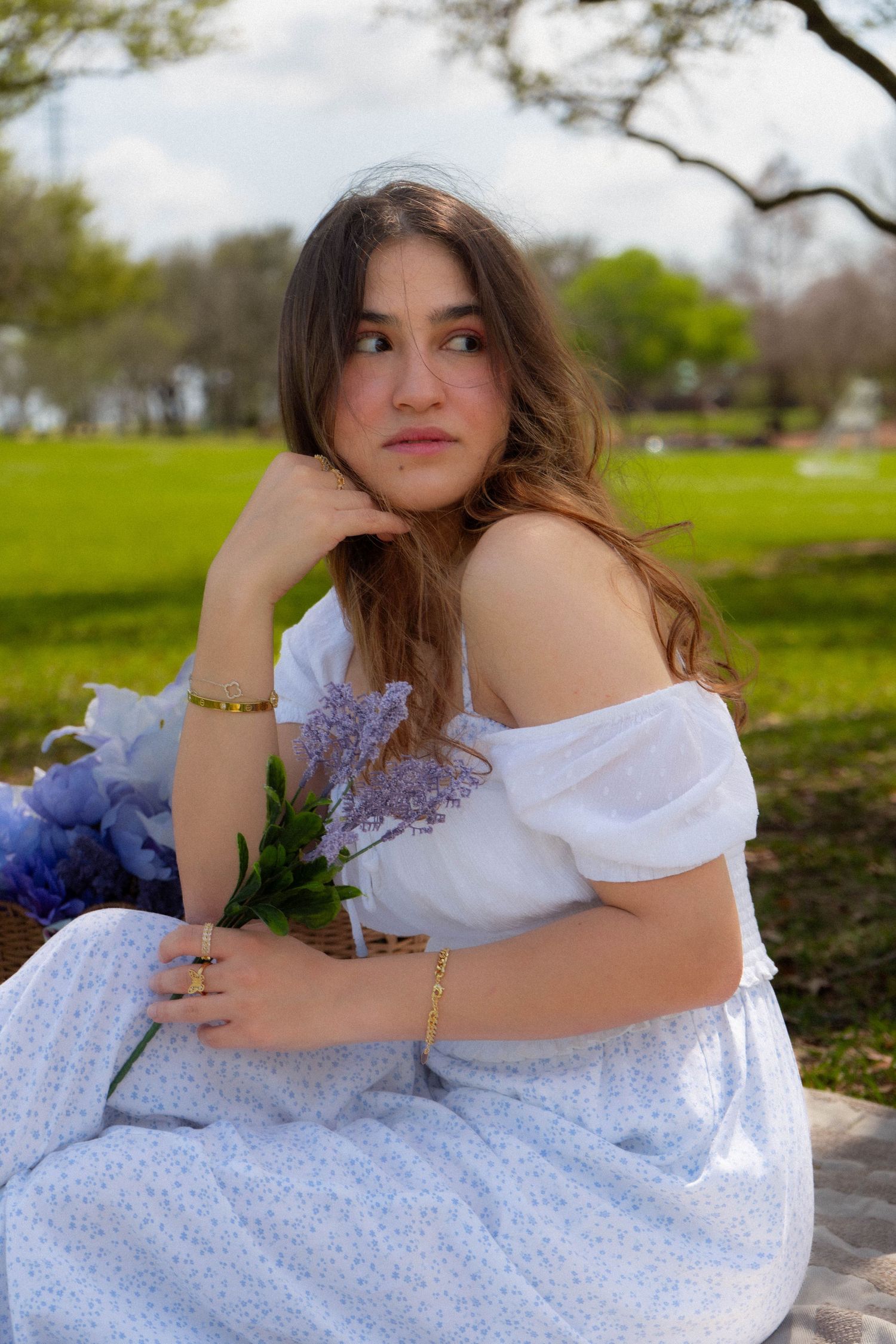 Young woman in white off-shoulder dress sitting in a park holding purple flowers in a dreamy outdoor portrait.