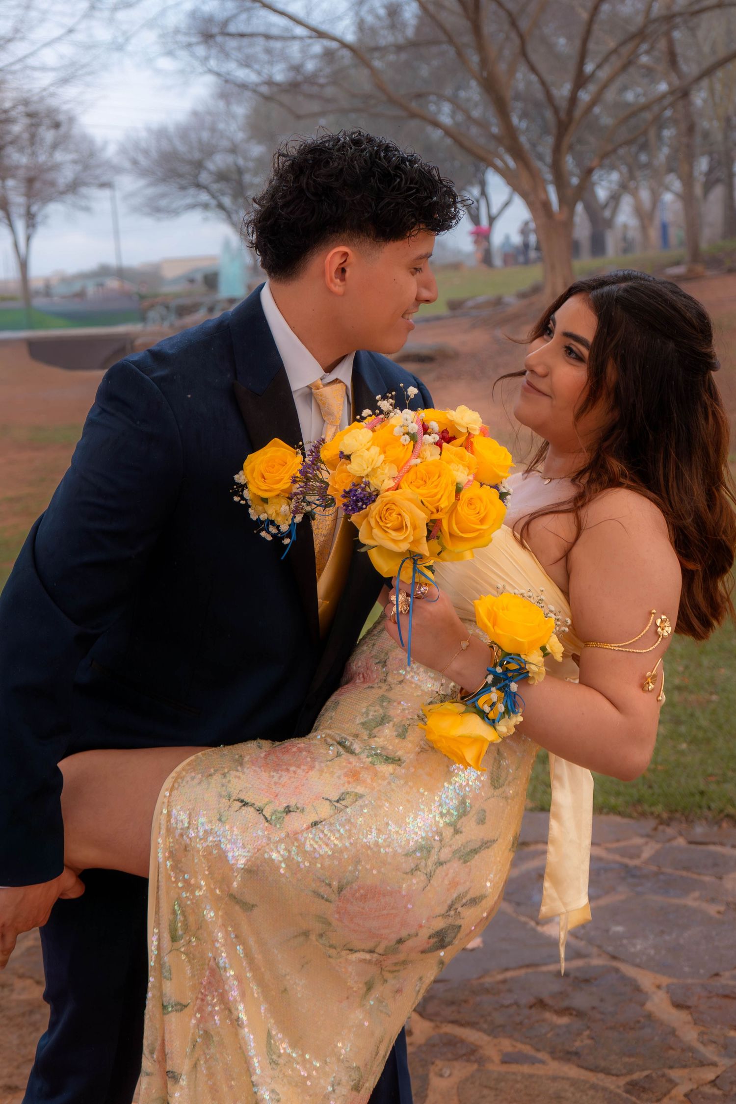 Prom couple sharing a romantic moment as young man lifts girl in gold sequin gown while holding yellow rose bouquet outdoors.