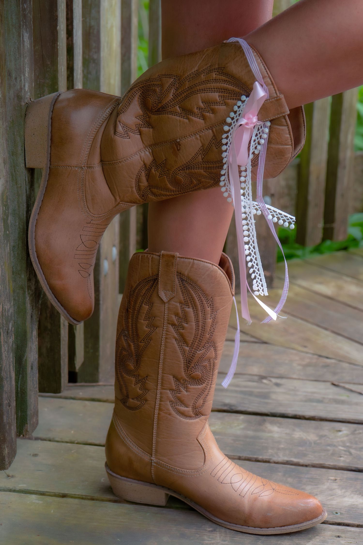 Brown cowboy boots with rhinestone and pink ribbon garter on wooden porch.