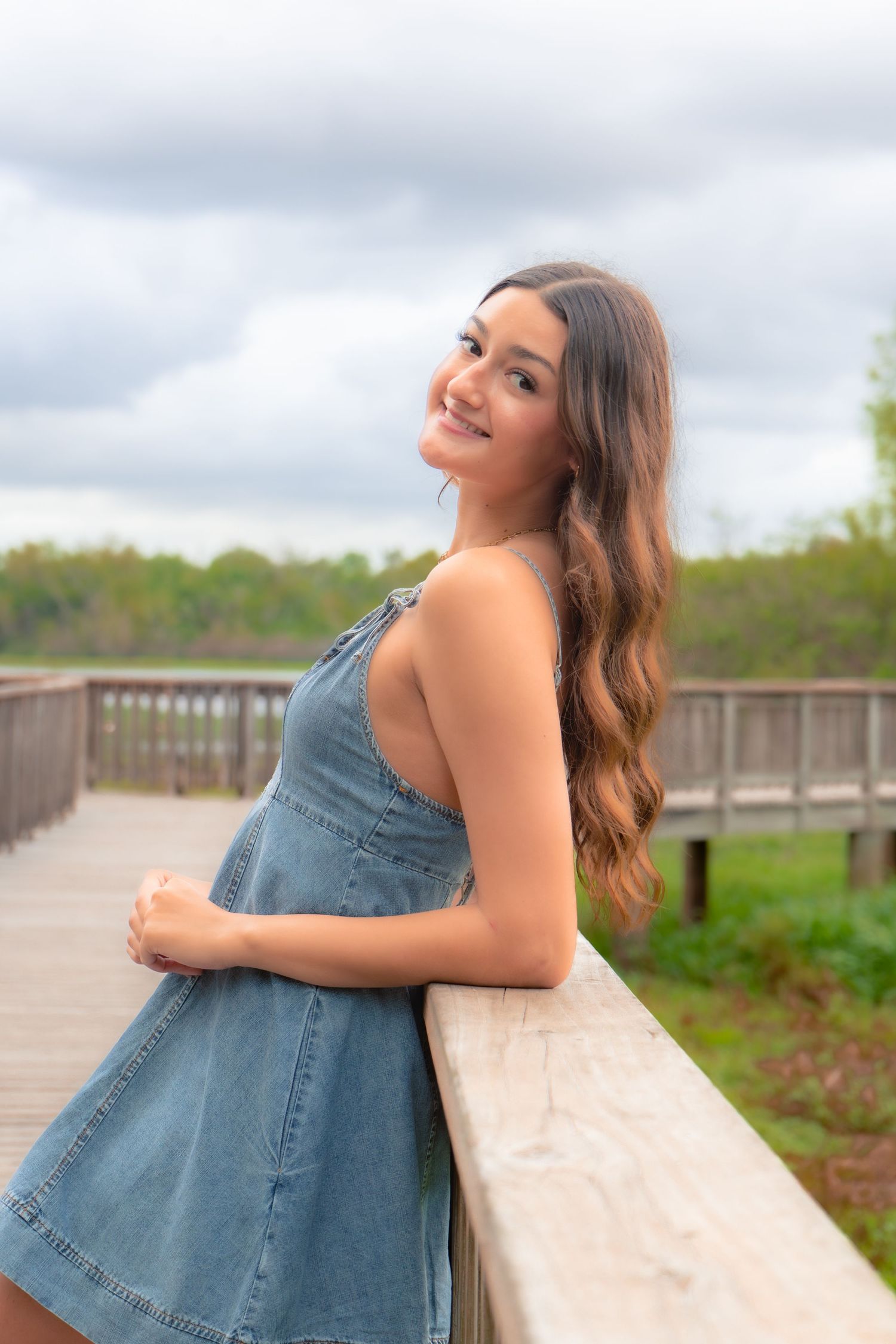 Smiling teenage girl in denim dress leaning on wooden railing outdoors.
