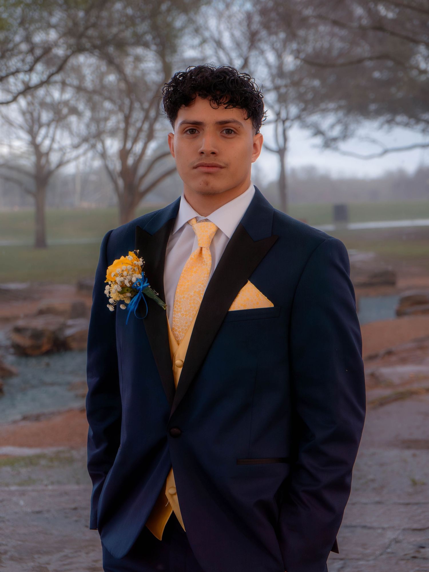 Young man in navy tuxedo with yellow tie and pocket square wearing yellow rose boutonniere posing outdoors for prom photos.