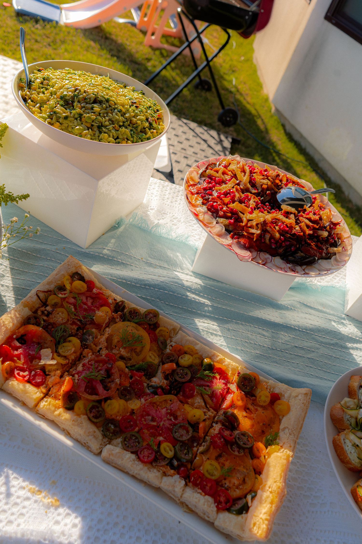Outdoor party food spread featuring a vegetable flatbread tart, grain salad, and colorful rice dish on a white tablecloth.
