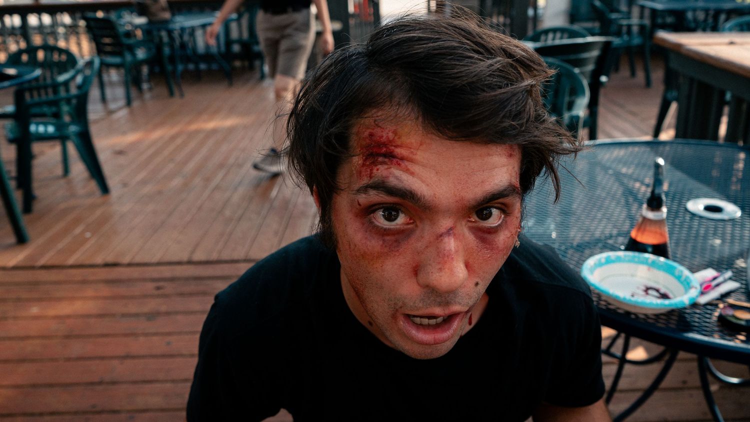 Close-up of a man with stage makeup cuts and bruises on his face at an outdoor patio.