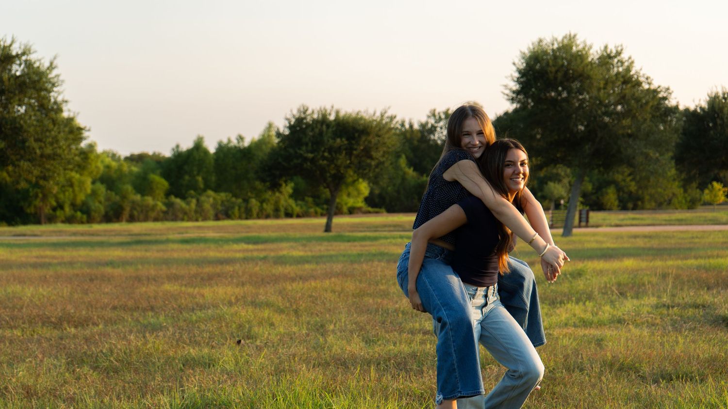 Two young women laughing outdoors, one giving the other a piggyback ride in a sunny green field.