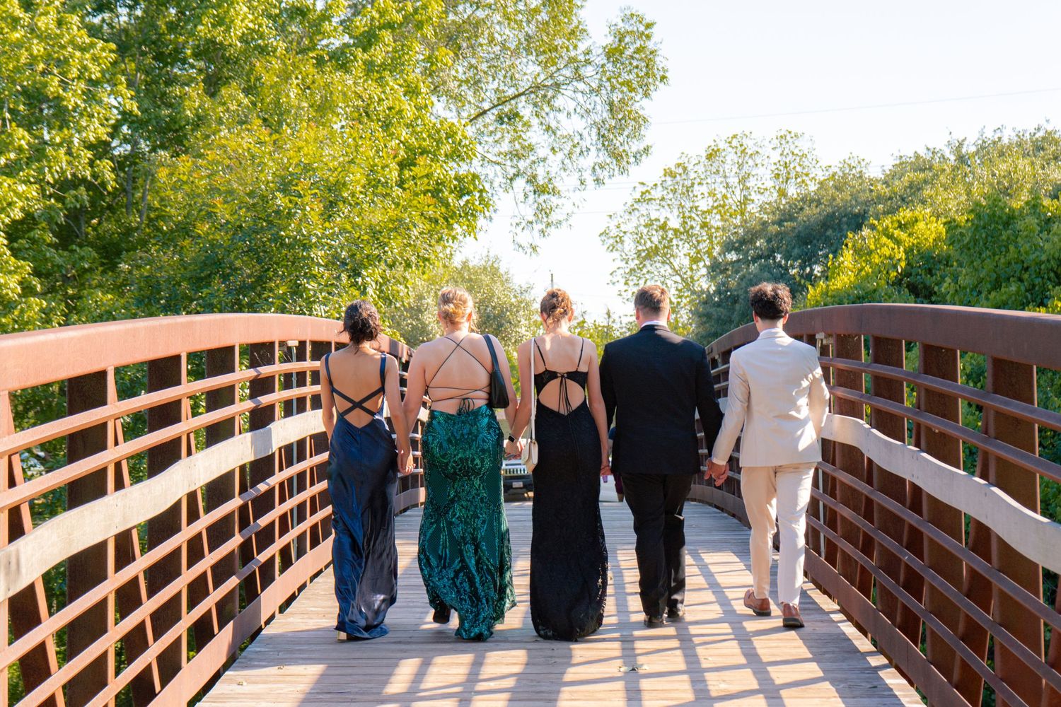 Group of prom-dressed teens walk away on wooden bridge surrounded by lush green trees.