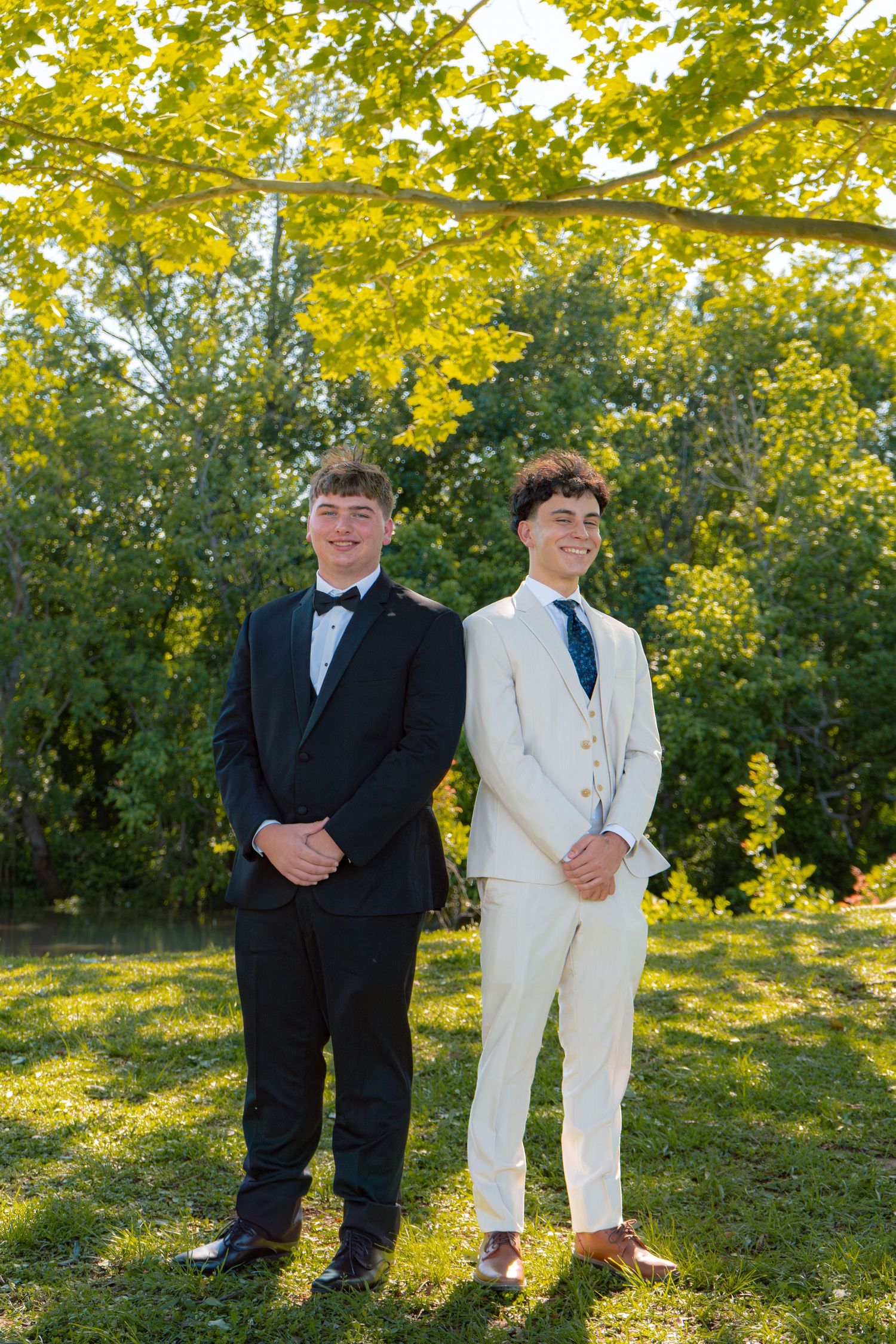 Two young men in formal suits, one black tuxedo and one cream suit, stand smiling outdoors under bright green trees.