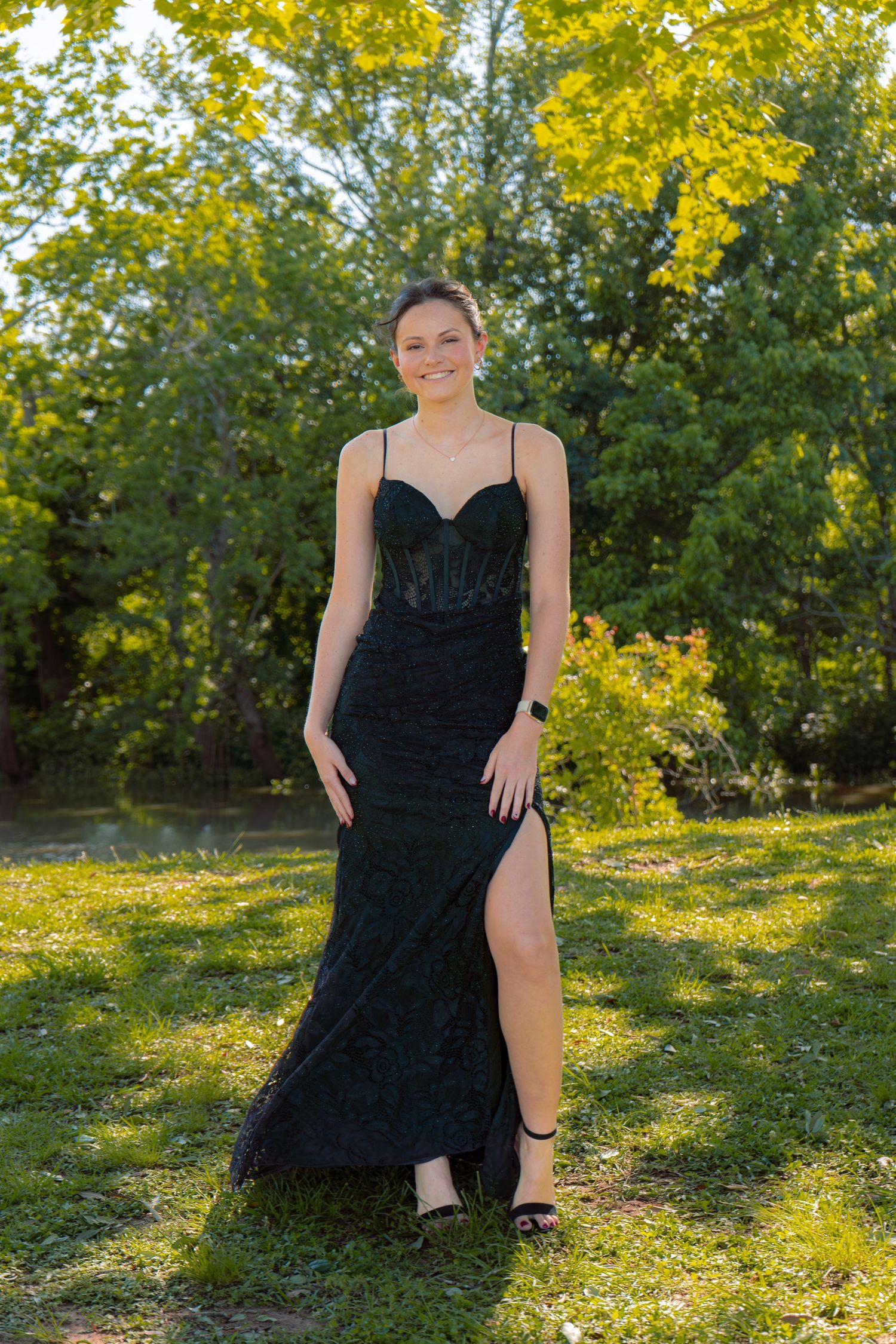 Young woman in elegant black sleeveless formal gown smiles while posing outdoors in sunlit green park.