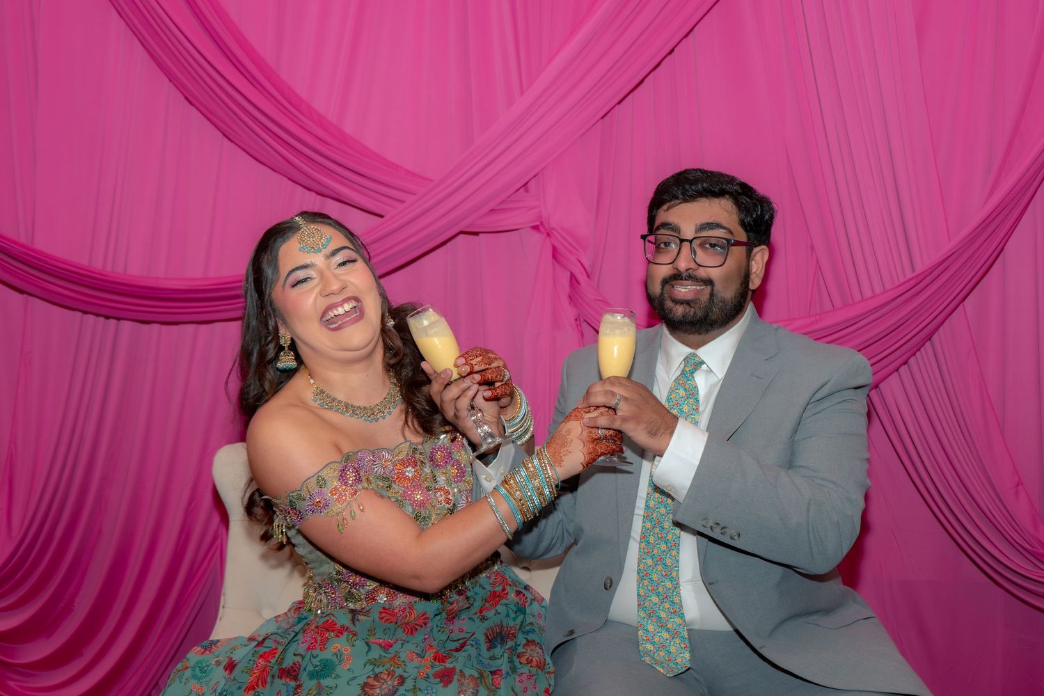 South Asian couple toasting with drinks, laughing at a pink draped celebration event.