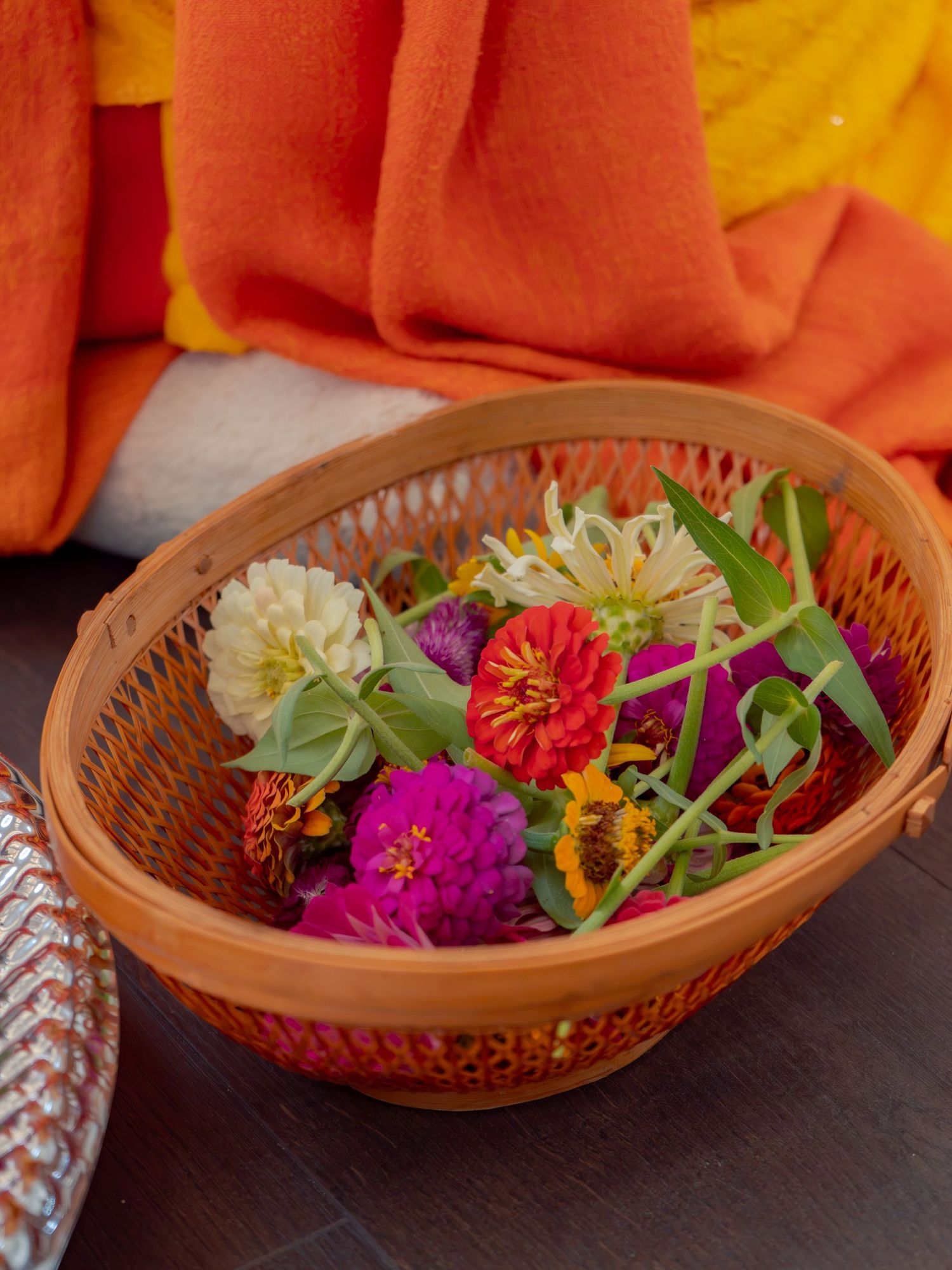 Colorful zinnia flowers arranged in a woven orange basket, placed near orange fabric offerings.