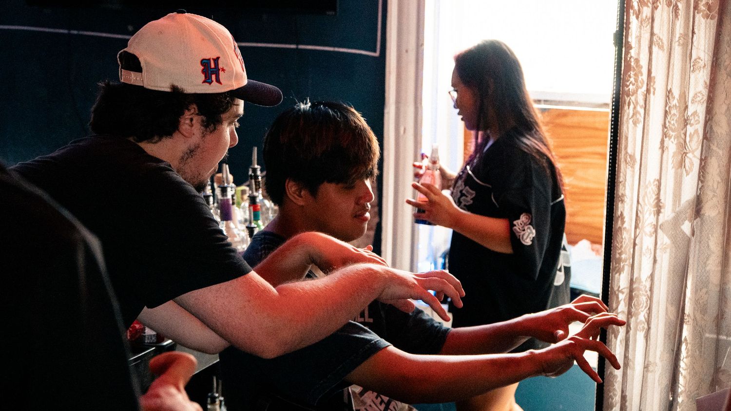Three young people gathered indoors, two leaning over a surface while a woman stands nearby in a casual setting.