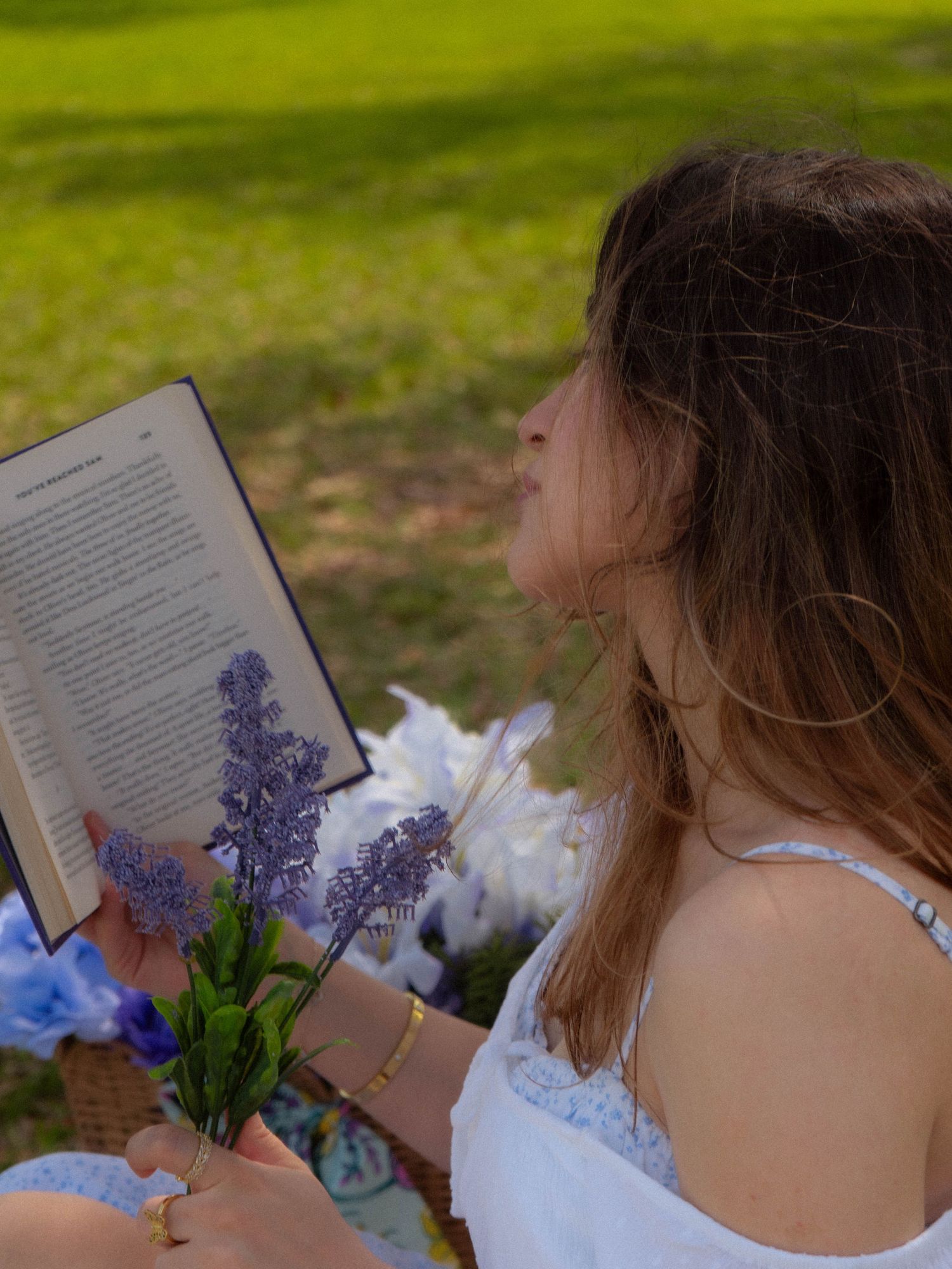 Young woman in white dress reading book while holding purple lavender flowers during outdoor picnic.