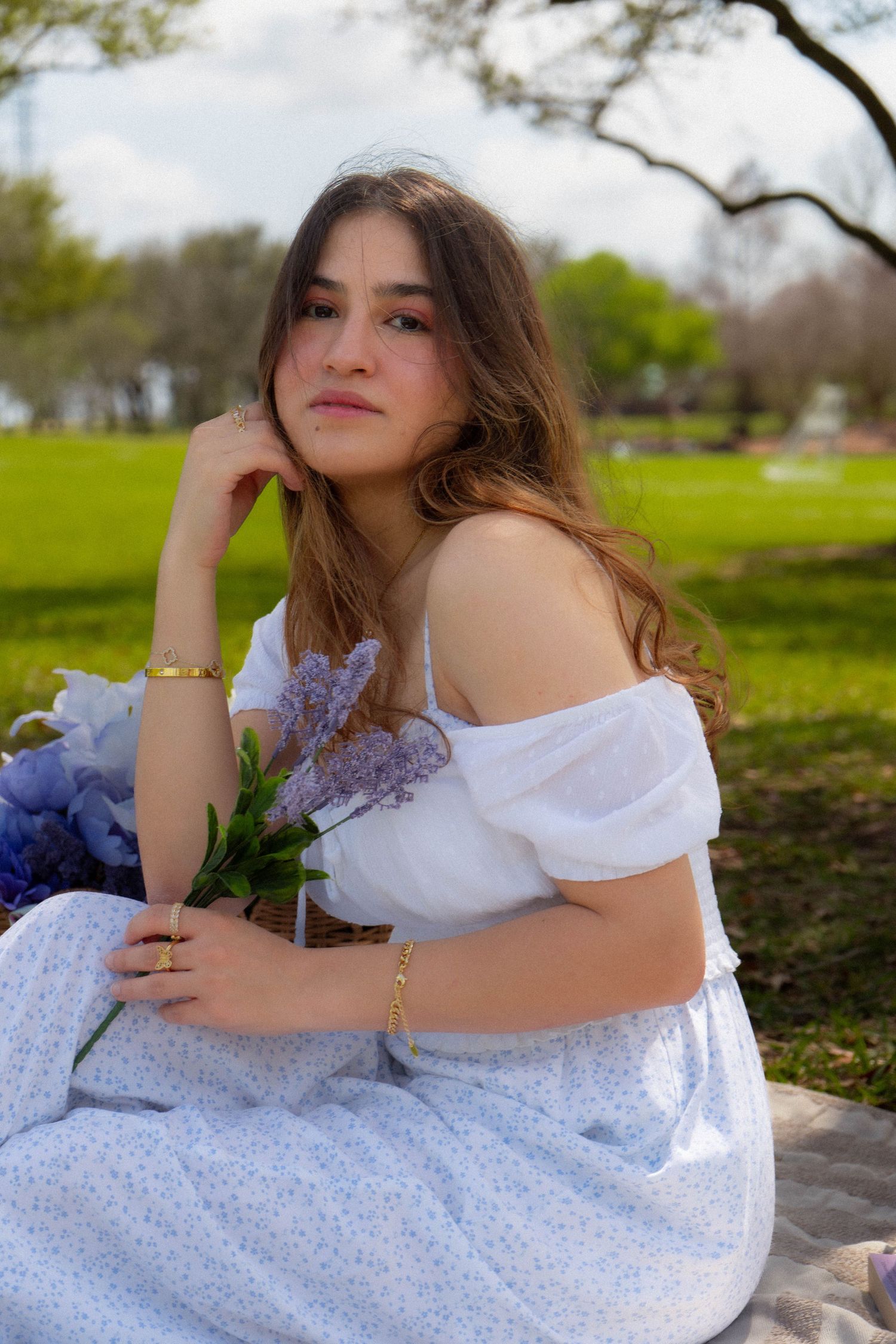 Young woman in white off-shoulder dress sitting in park, holding lavender flowers in wicker basket.