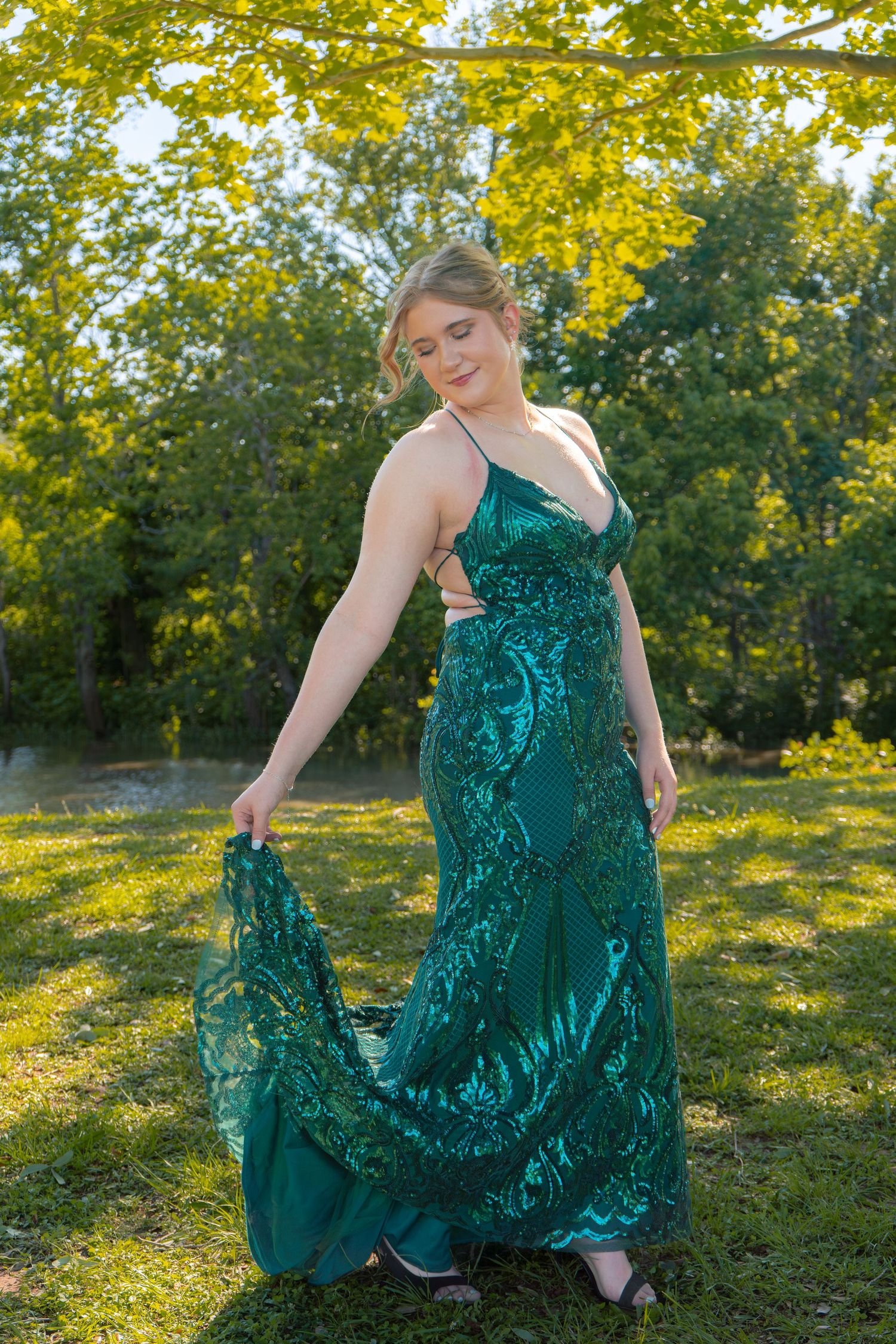 Young woman in green sequin prom gown poses outdoors in sunlit park.