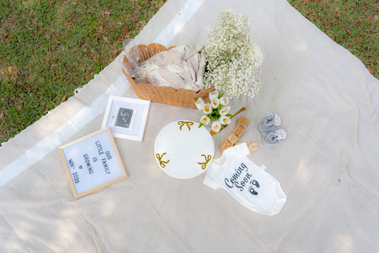 Aerial view of a baby shower flat lay with a onesie, floral arrangement, picture frames, and wicker basket on white blanket.