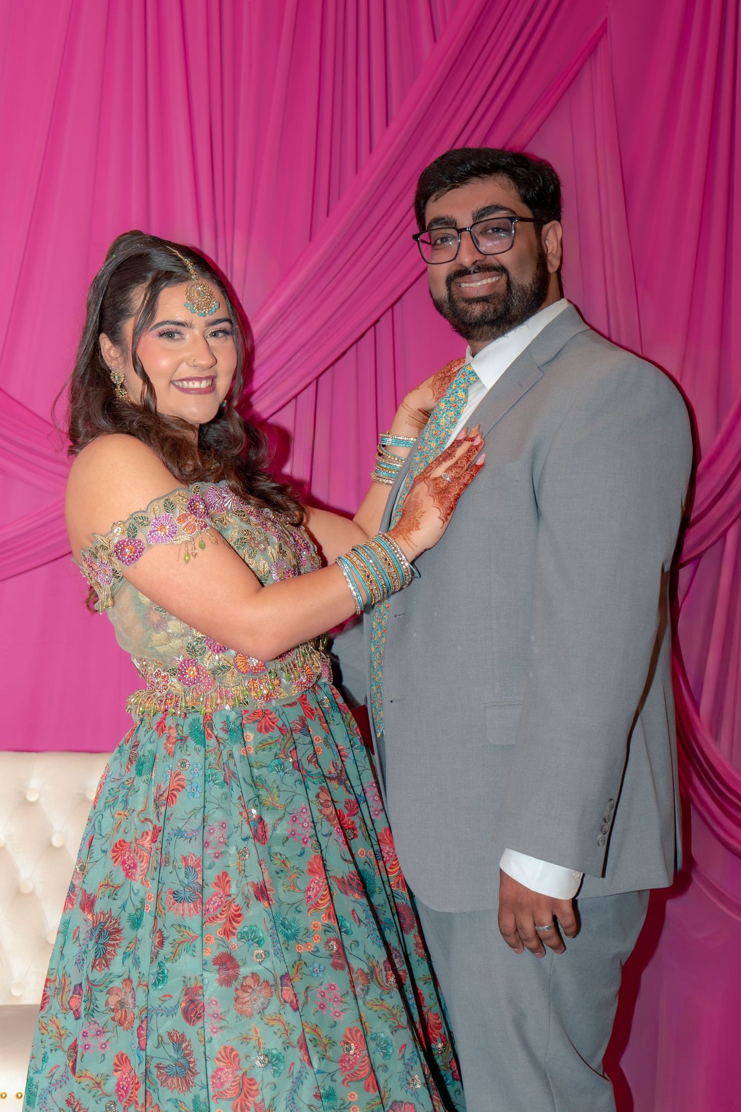 South Asian bride in teal floral lehenga adjusting grooms grey suit lapel before pink drape at wedding celebration.
